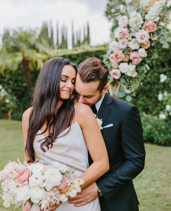 A couple, a woman in a white dress and a man in a suit, embrace outdoors next to a bouquet of pink and white flowers, with a floral arrangement in the background.