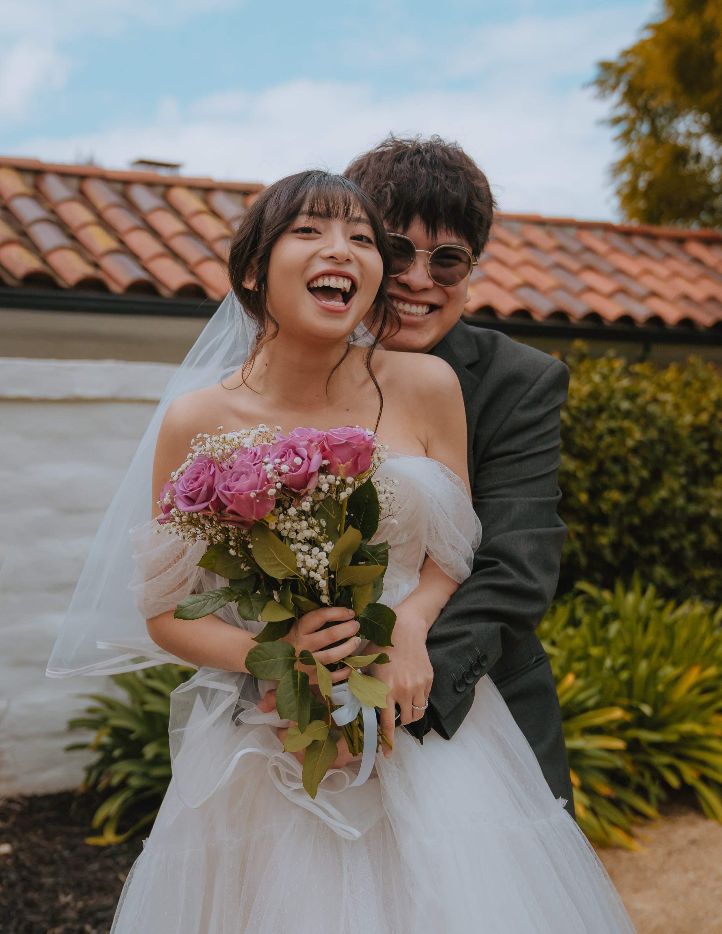 Joyful bride holding a bouquet of pink roses and wearing a wedding dress, hugging groom dressed in a black suit with sunglasses, outdoors during daytime.