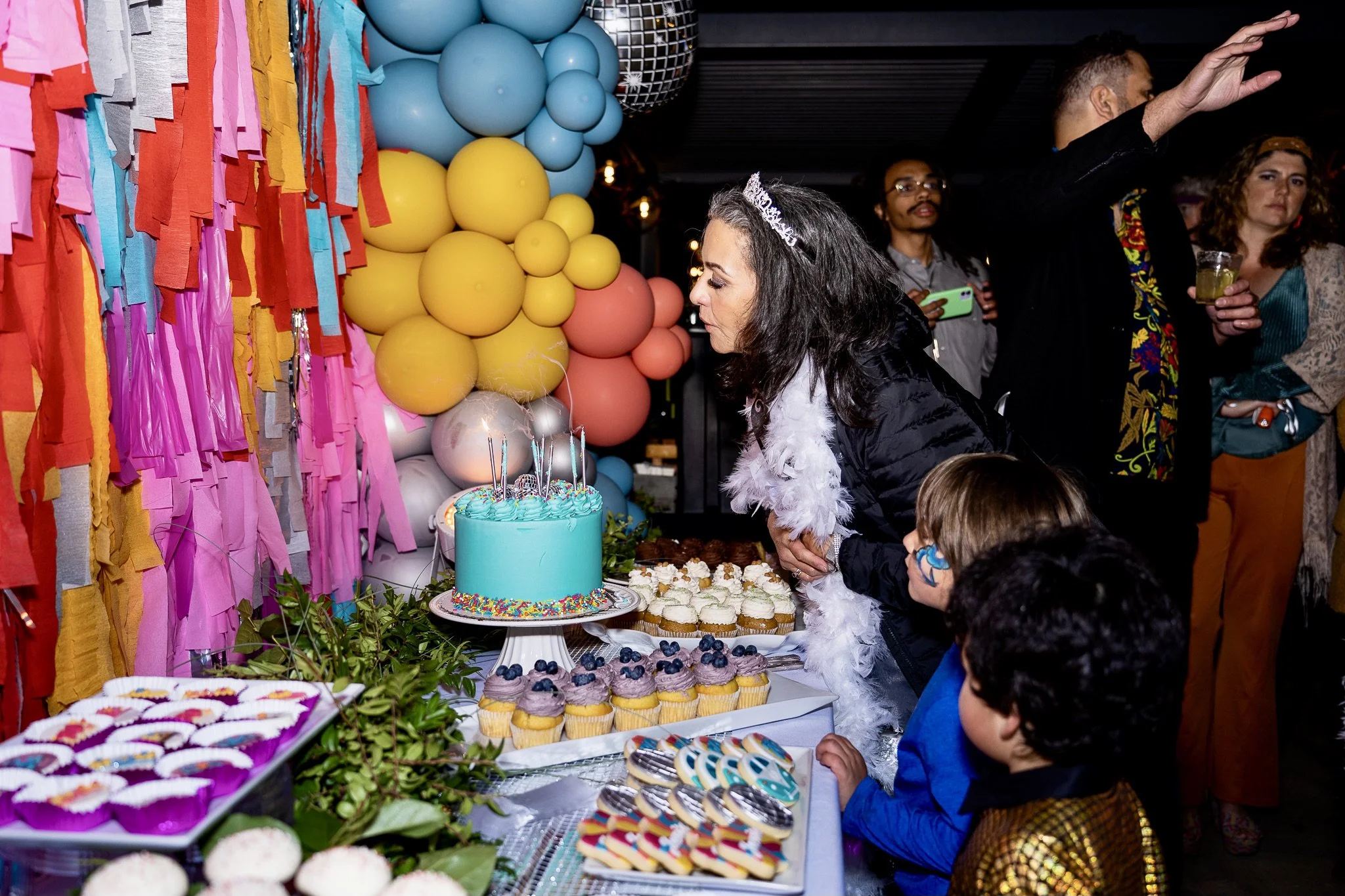 A woman wearing a tiara and feather boa is blowing out candles on a birthday cake at a party with colorful balloons and cupcakes. Other people are in the background, some holding drinks and taking photos.