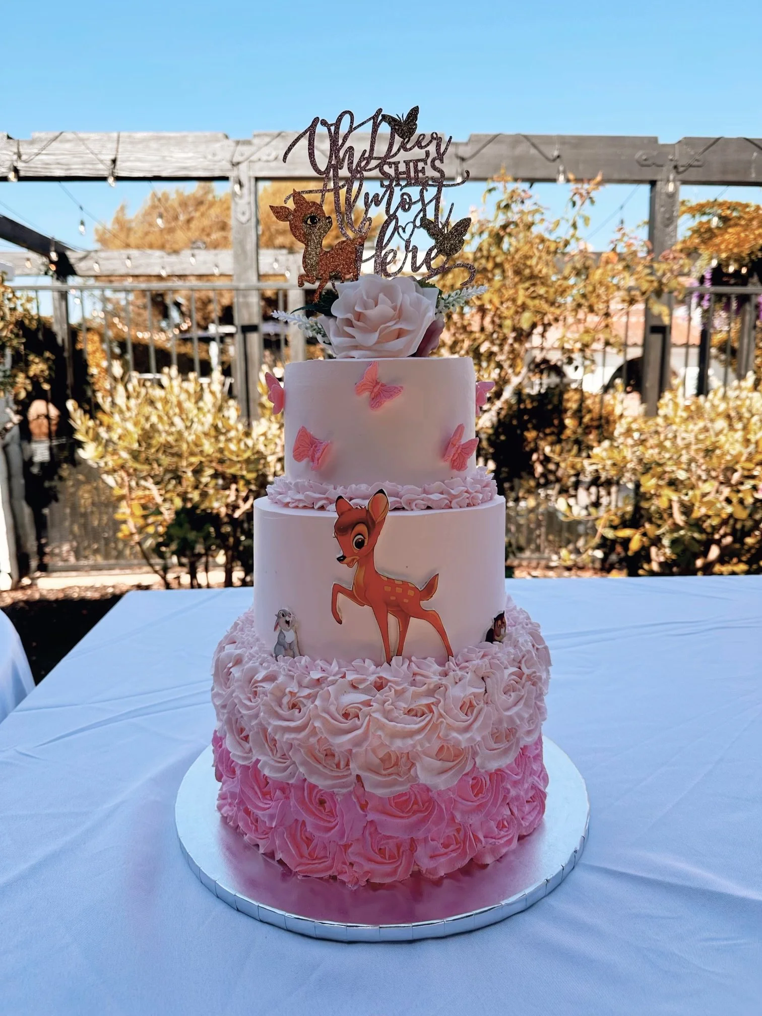Pink and white tiered birthday cake with butterfly and deer decorations on a table outdoors.
