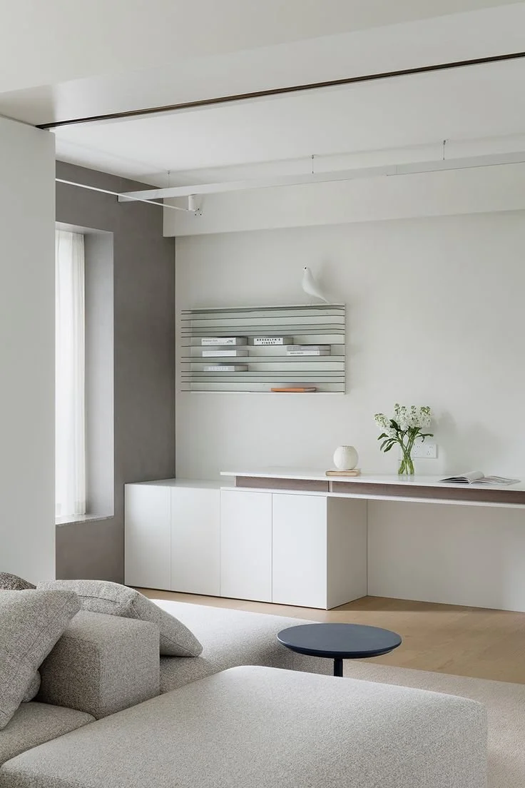 Minimalist living room with a light-colored sofa, a small round black side table, and a white built-in cabinet with decorative items on top, including a vase with flowers and a book. There is a window with sheer curtains, and a modern wall shelf with books and a bird sculpture on the wall.