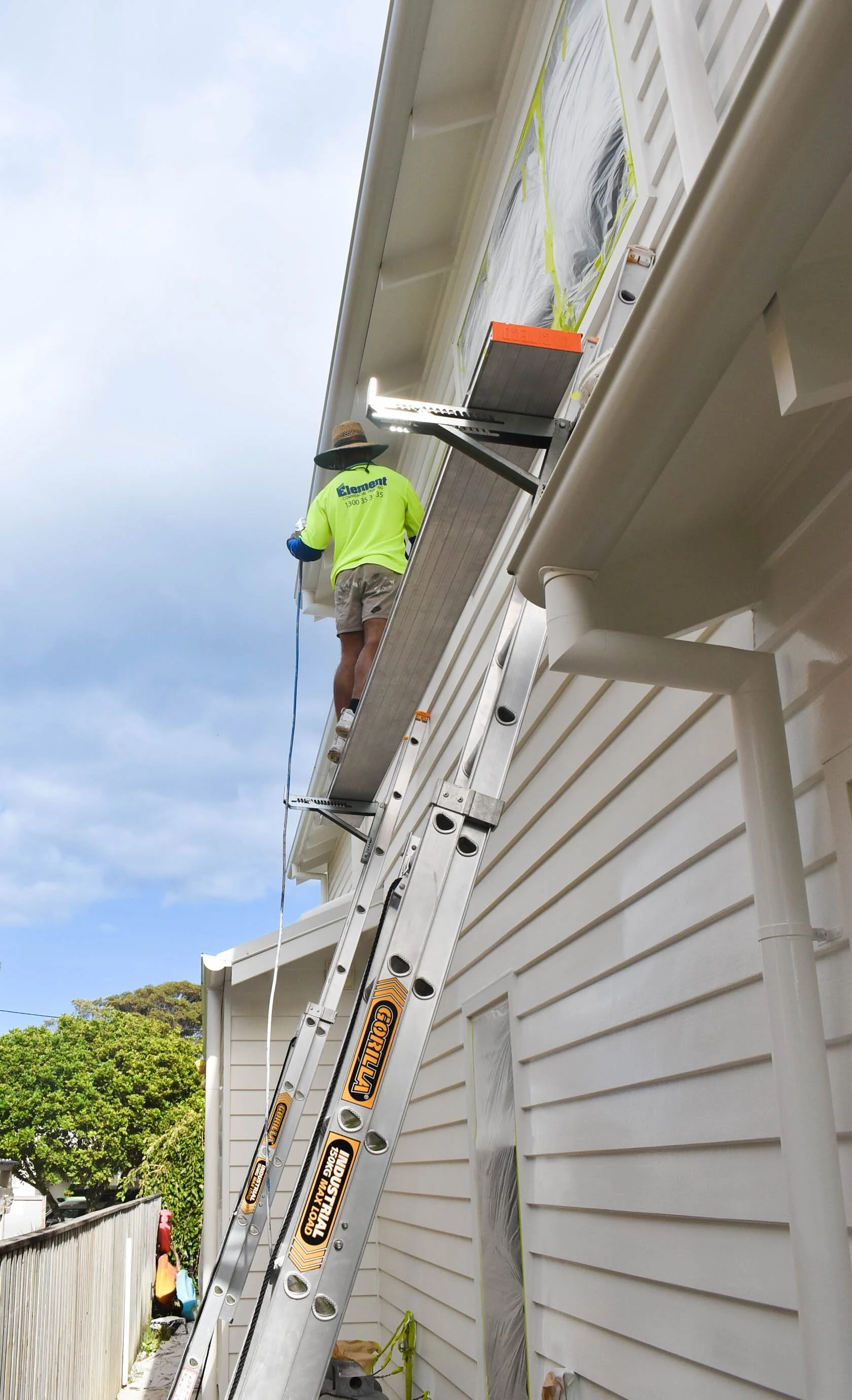 A person wearing a green shirt and shorts standing on an extension ladder leaning against a house, working on the roof or gutter. The house has white siding, and the background shows blue sky and trees.