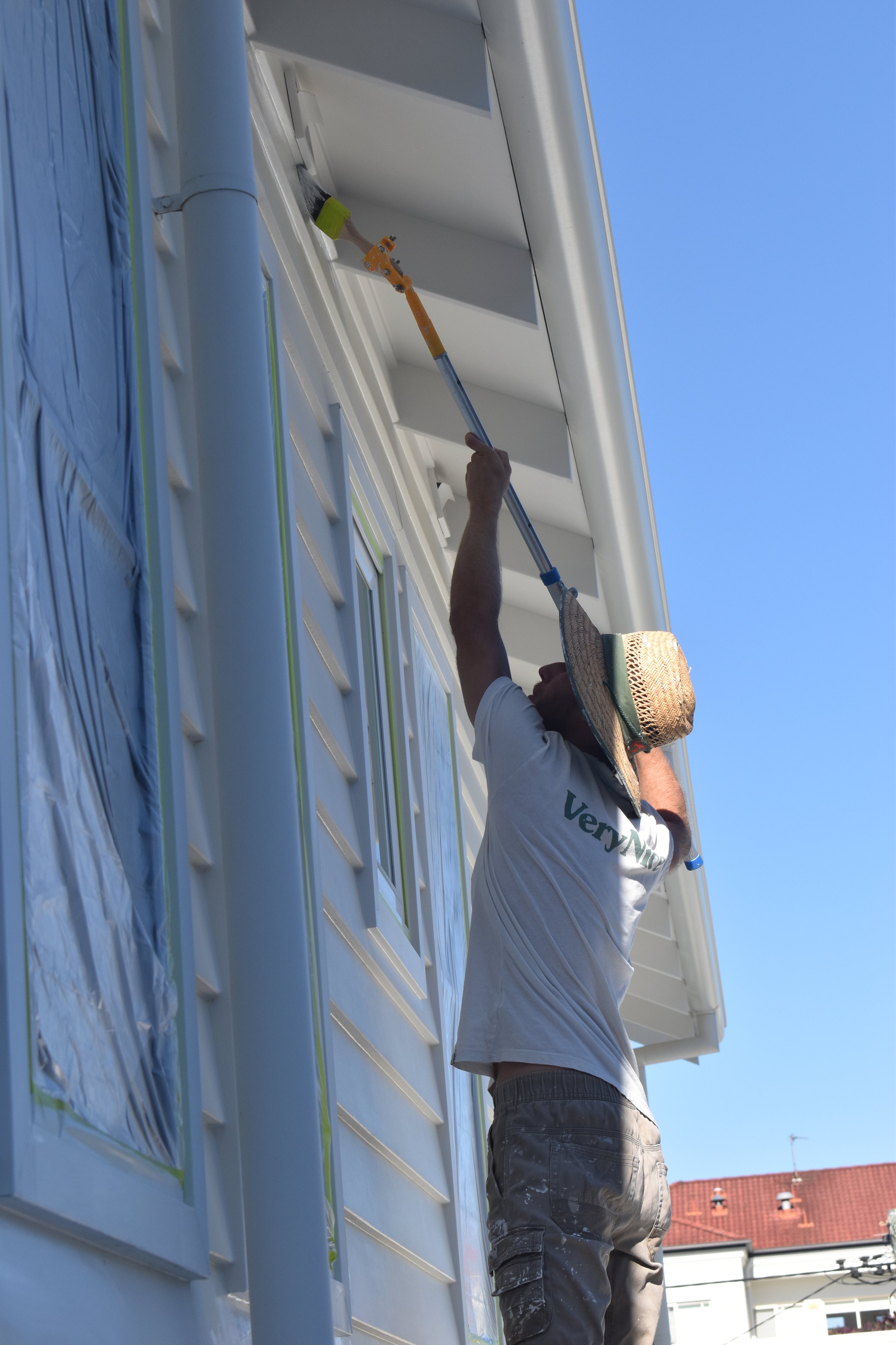 A person painting the exterior of a house with a roller on an extension pole under a clear blue sky. The individual is wearing a wide-brimmed hat and casual work clothes.