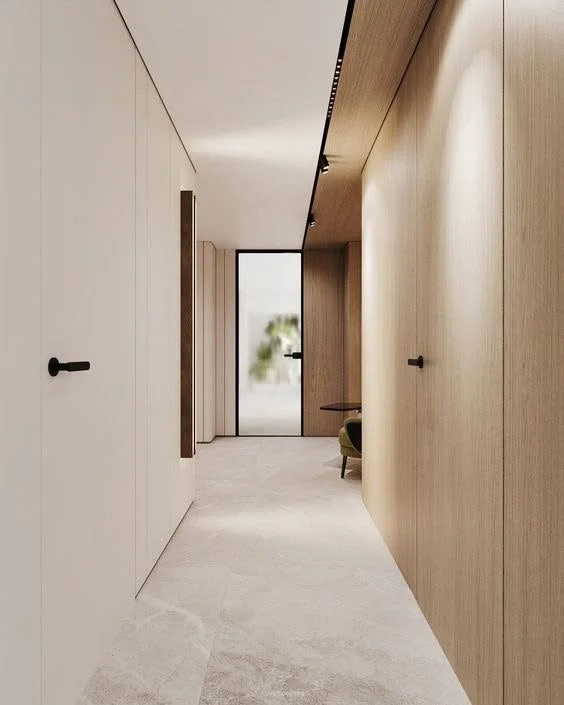 Modern minimalist hallway with light wood walls, white ceiling, and a gray floor. Black door handles and a glass door at the end with blurry outdoor view.