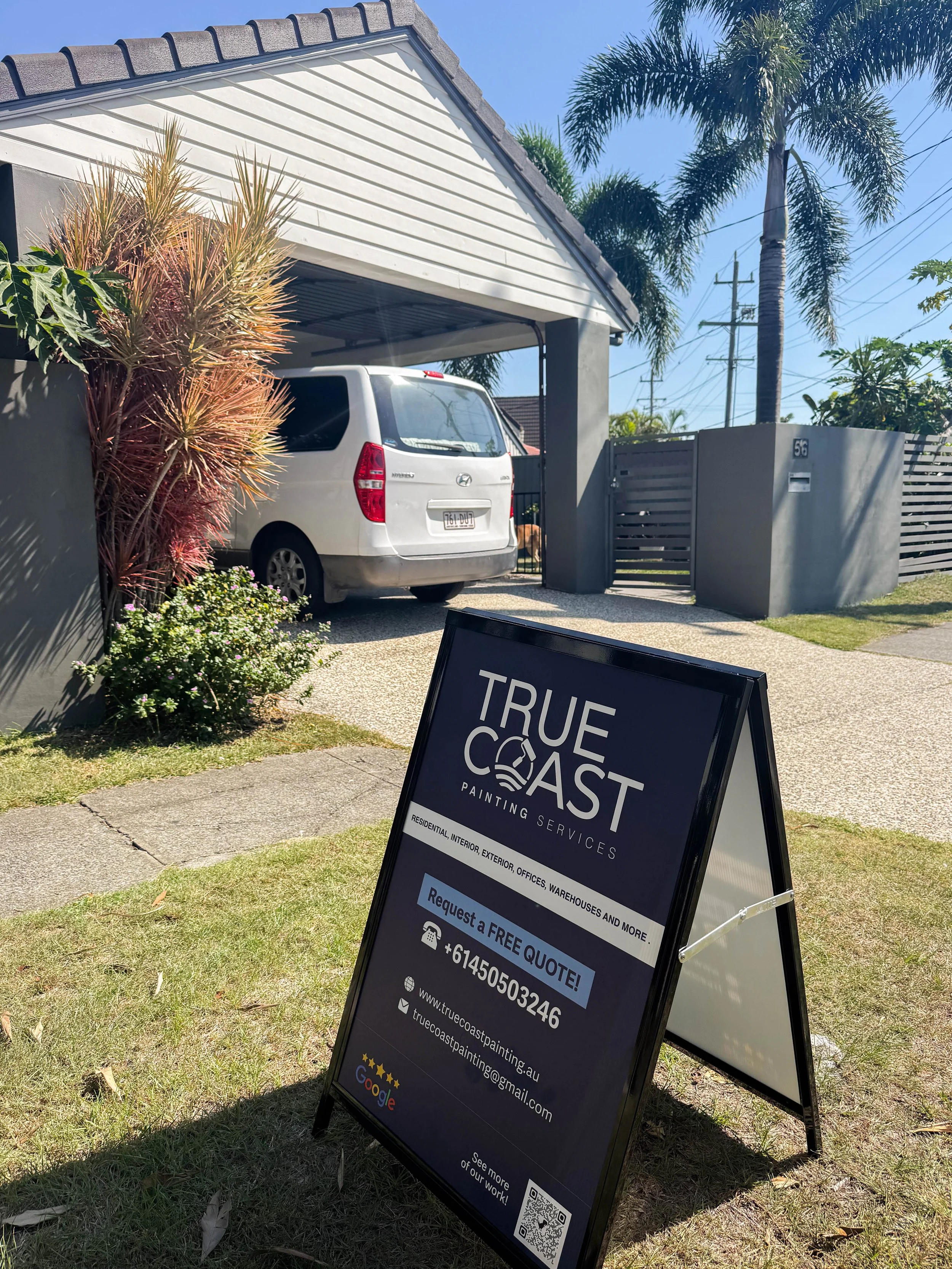 A black sidewalk sign for True Coast Painting Services placed on a grassy lawn in front of a house with a carport, white minivan, and tropical plants, advertising free quotes and contact information.