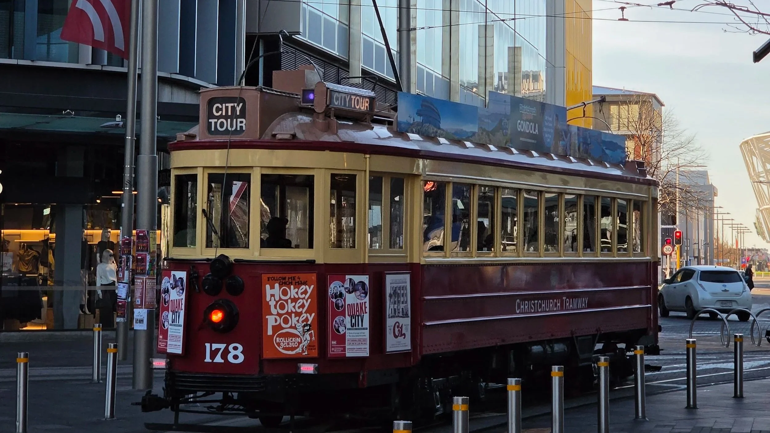 A vintage Christchurch tram in red and cream colors on a city street, with advertisements and urban buildings in the background.