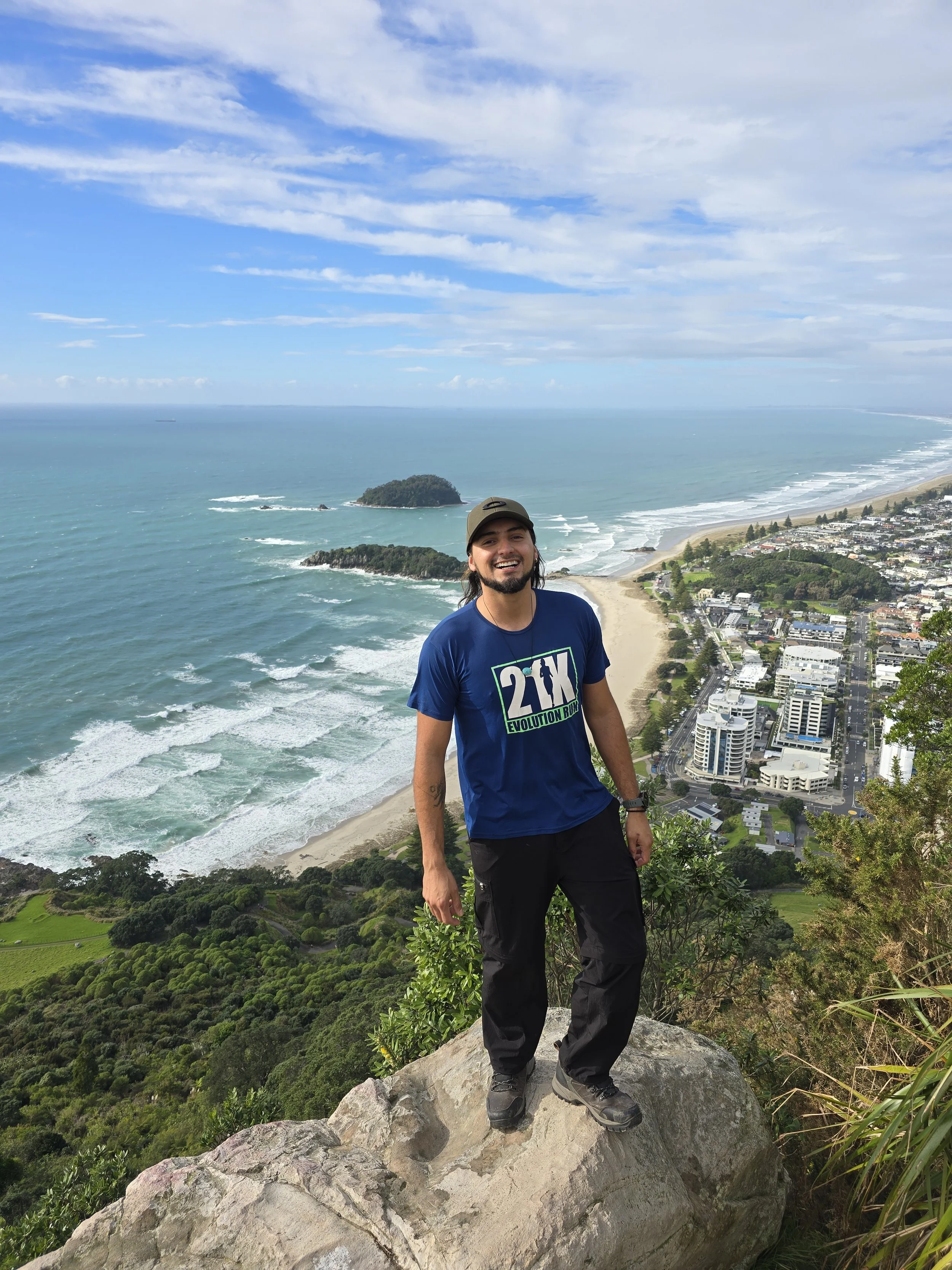 A man with long hair and a beard, wearing a blue t-shirt and black pants, smiling and standing on a large rock with a scenic coastal landscape in the background. The background features the ocean, a small island, and a city along the shoreline with cloudy sky overhead.