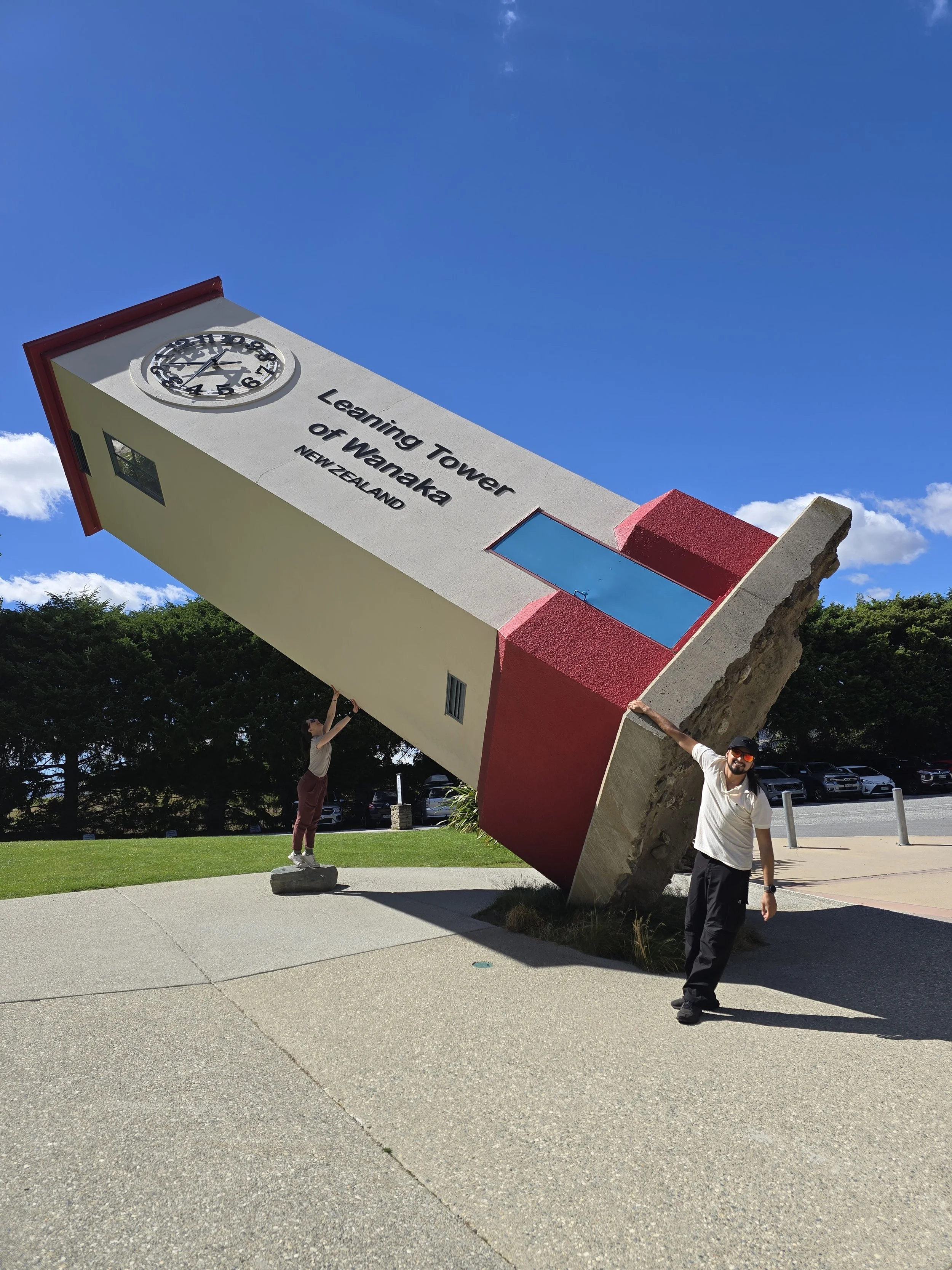 A large, tilted clock tower at Learning Tower of Wānaka in New Zealand with two people posing beside it under a clear blue sky.