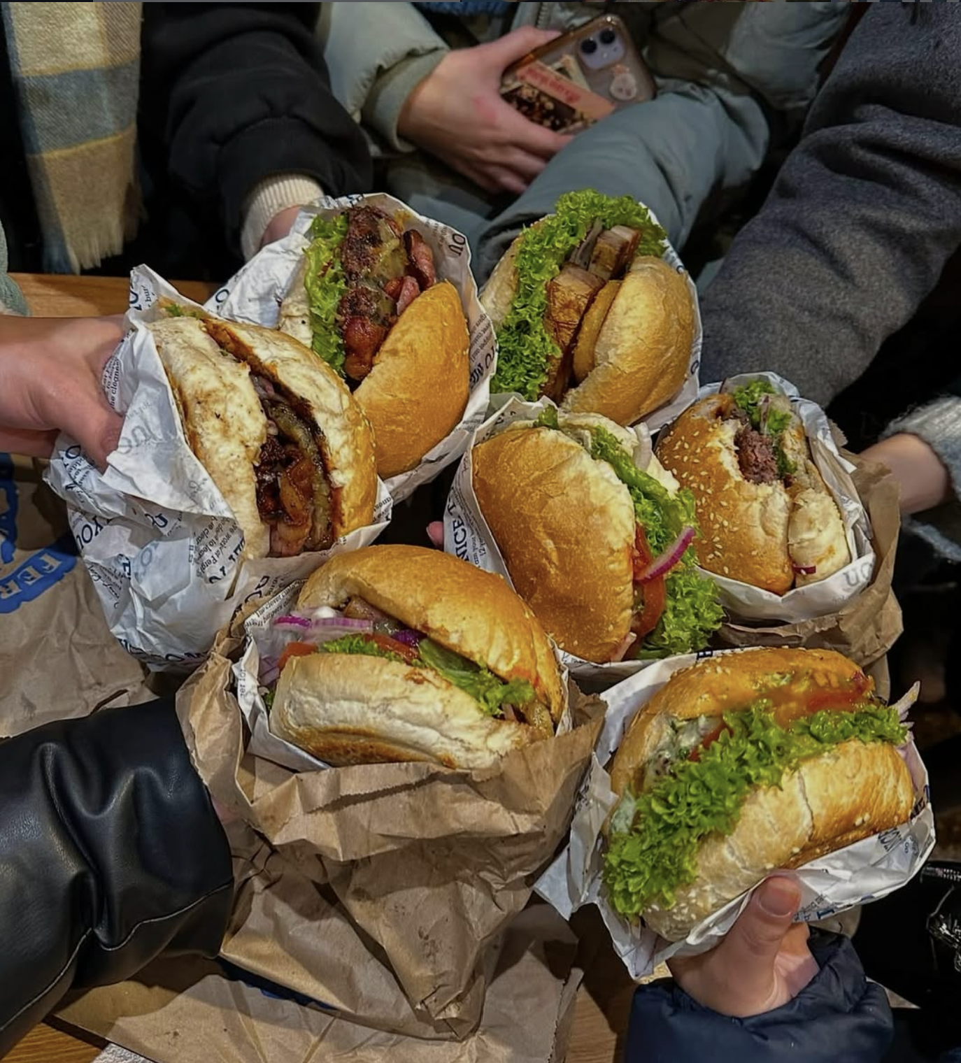 Several people holding and presenting various sandwiches with lettuce, tomatoes, and meats, wrapped in paper, over a table.