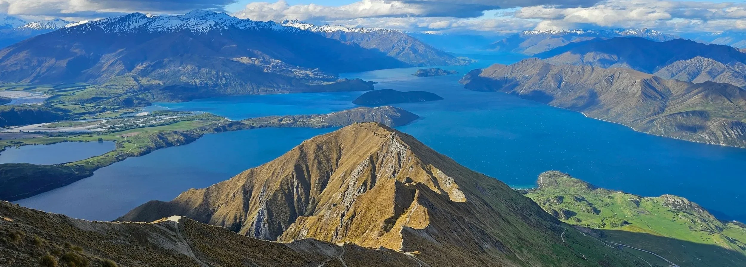Scenic view of a mountain range with snow-capped peaks in the background, several large lakes with blue water in the middle ground, and rolling green hills and cliffs in the foreground.