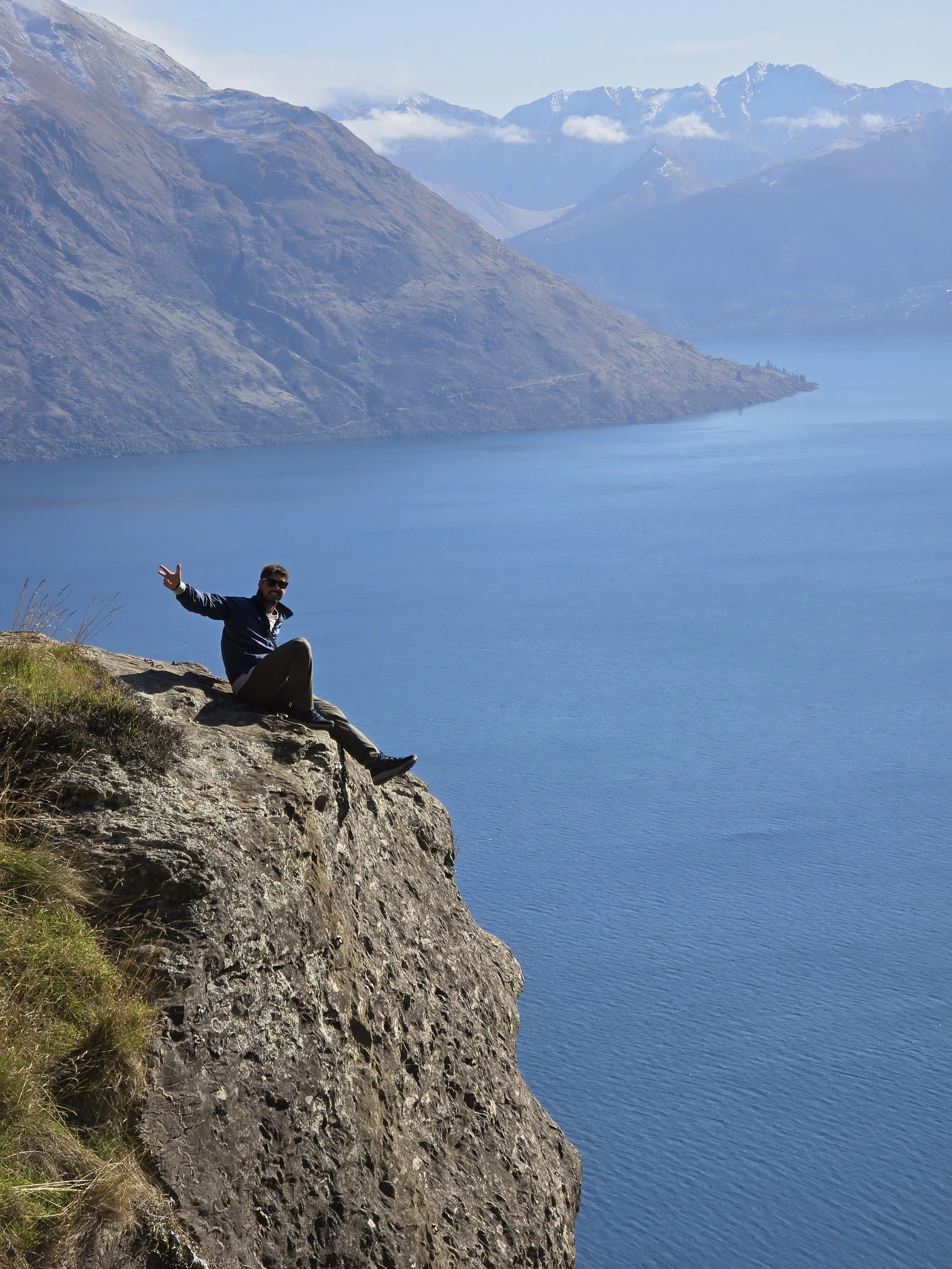 A person sitting on a rocky ledge next to a large body of water, with mountains in the background under a clear sky.