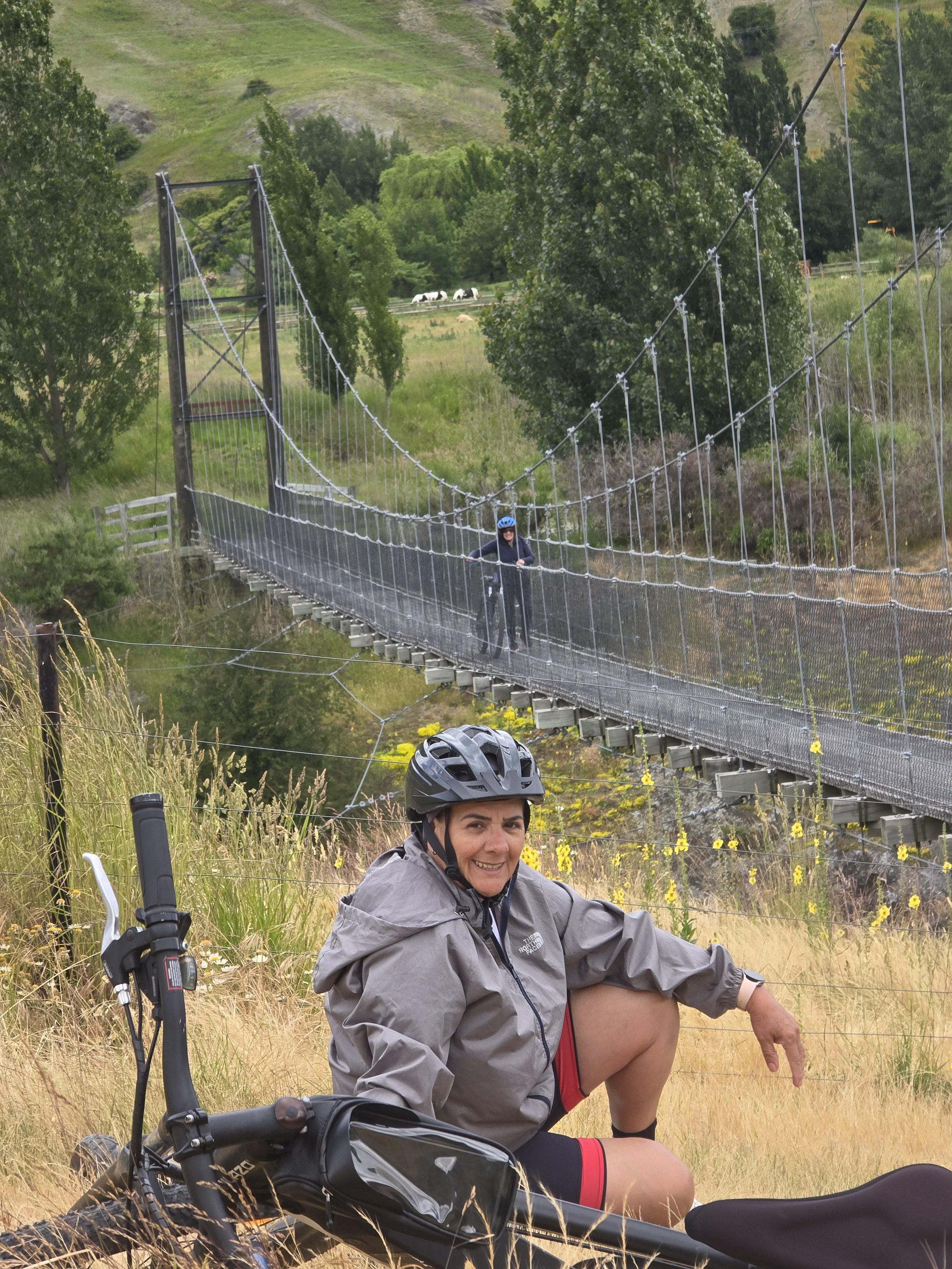 A woman sitting on the ground in a grassy field wearing a gray jacket and cycle helmet, smiling at the camera with mountain scenery and a suspension bridge in the background, and a bicycle in the foreground.