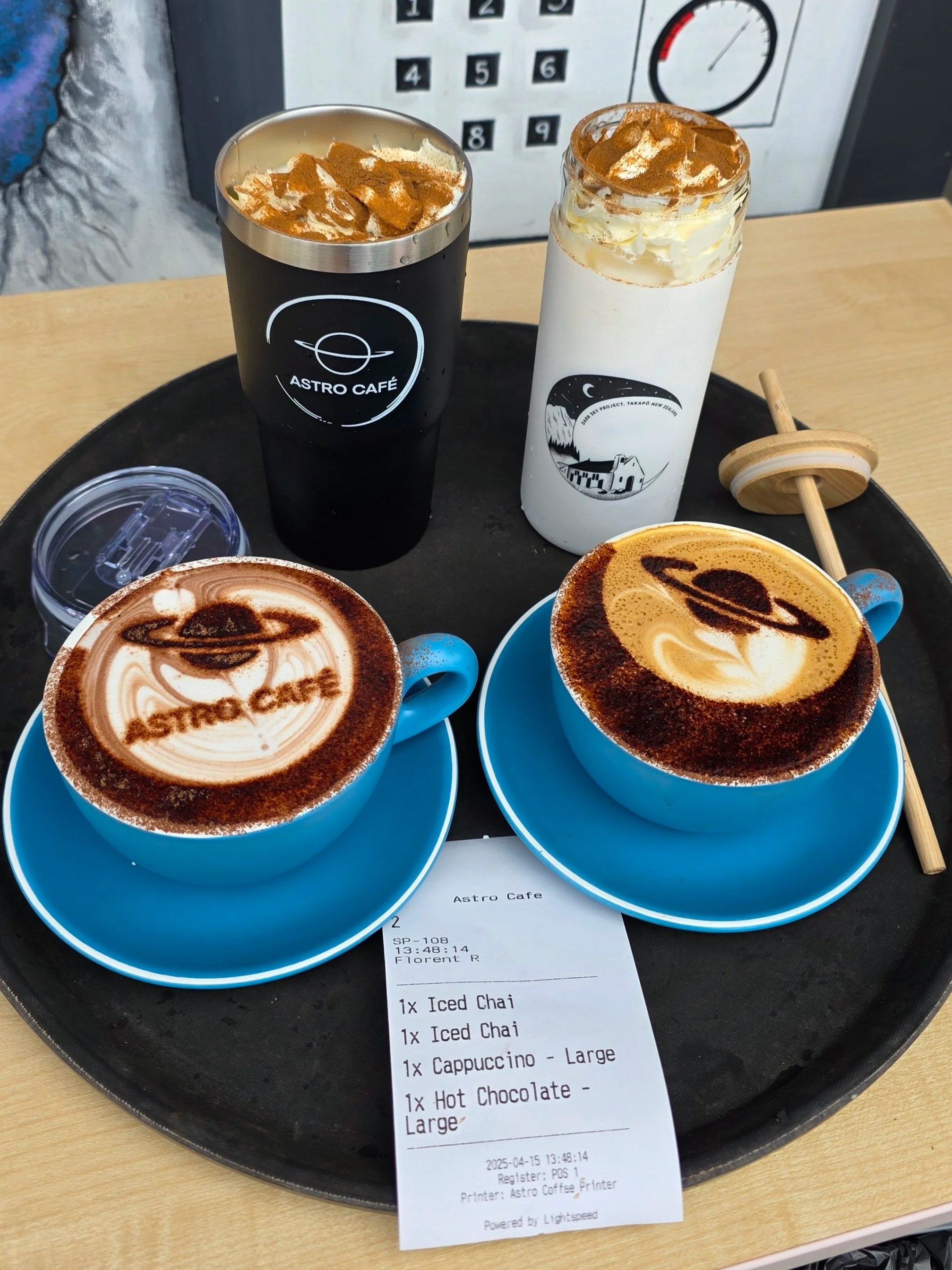 Two coffee drinks with latte art on blue cups, placed on a black tray, with a receipt listing iced chai, cappuccino, and hot chocolate, in a cafe setting.