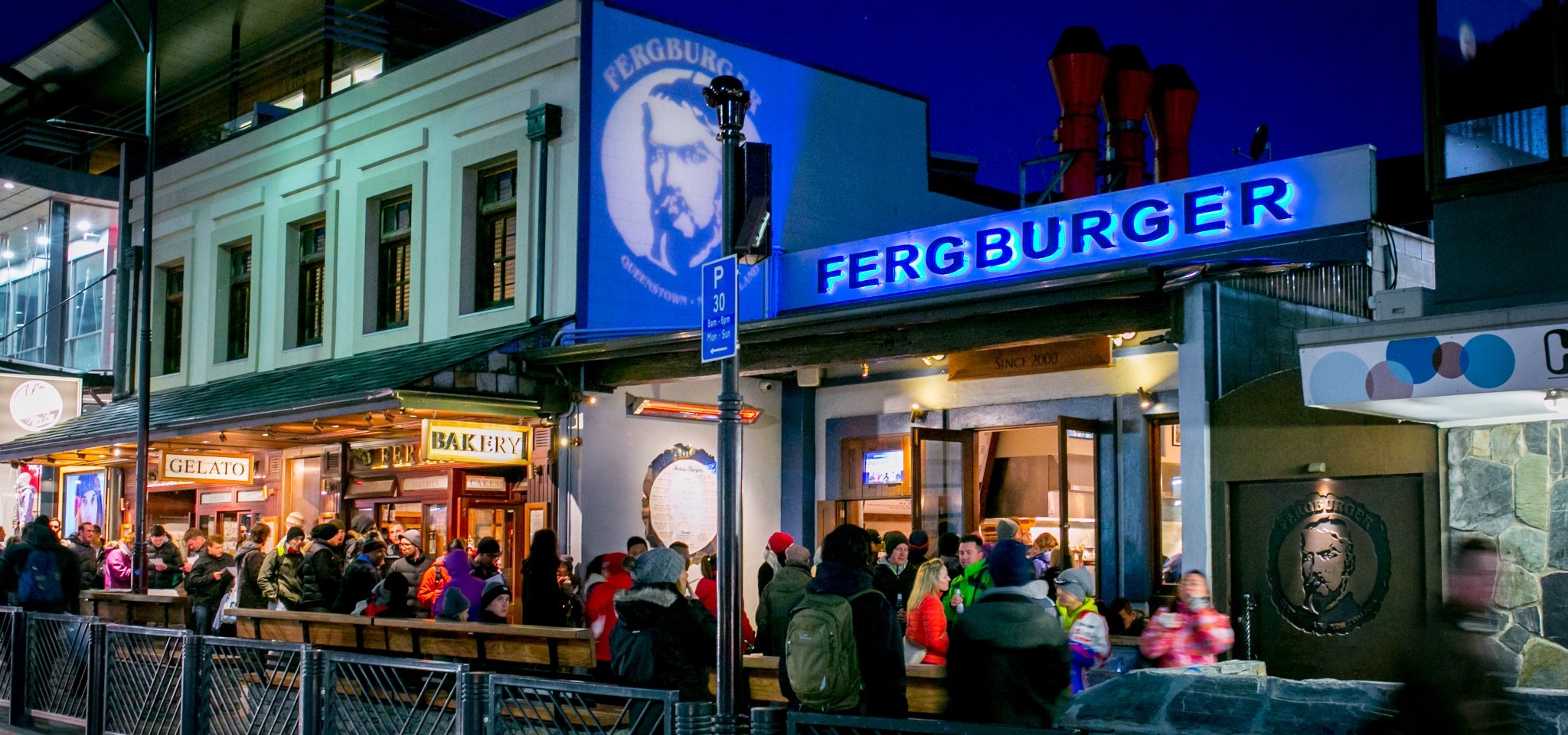 Night view of a busy street with a restaurant named Ferburger and a bakery, gelato shop, and outdoor seating, with a crowd of people.