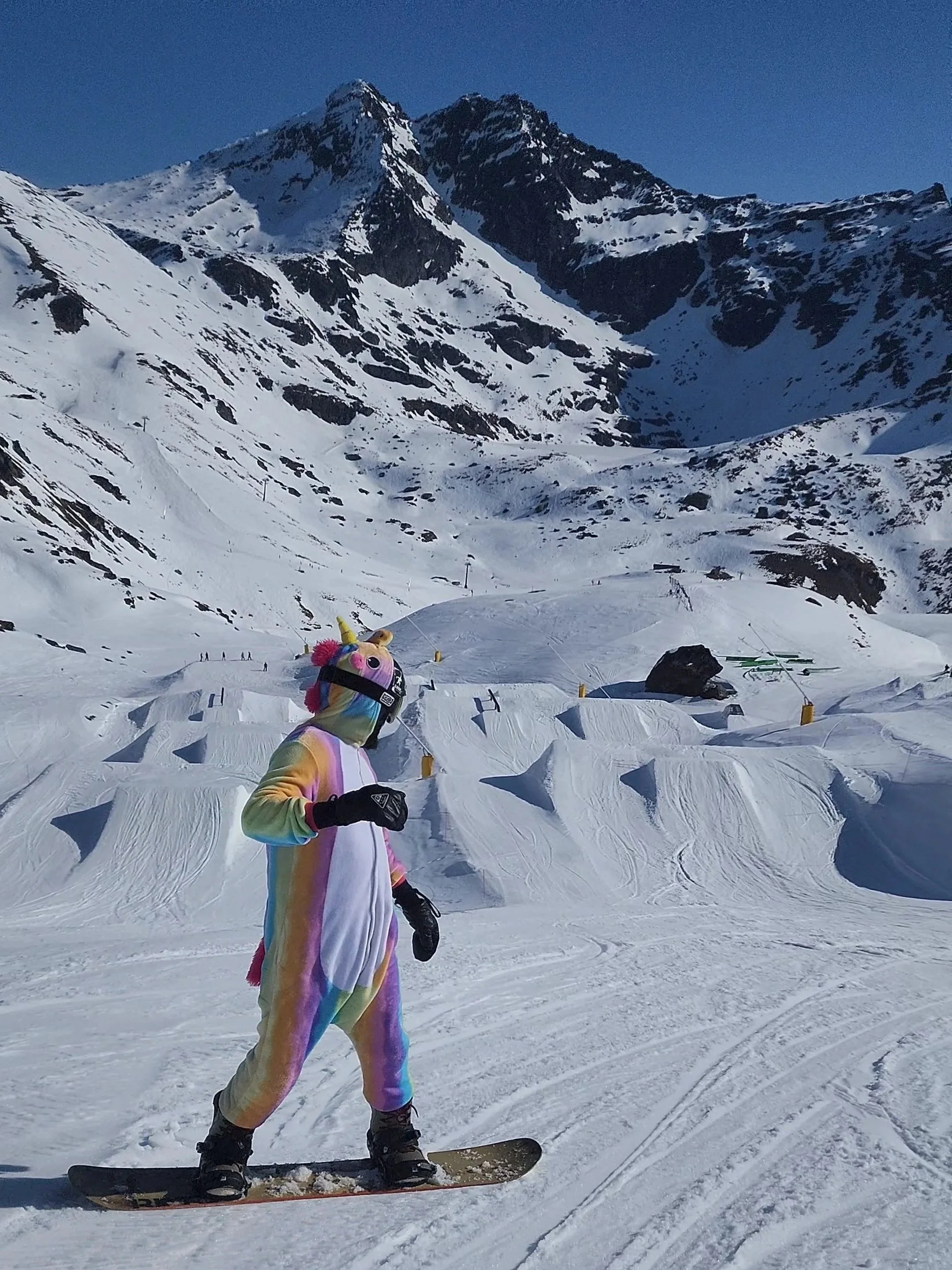 A person in a rainbow-colored unicorn onesie snowboarding on a snowy mountain slope with mountains in the background.