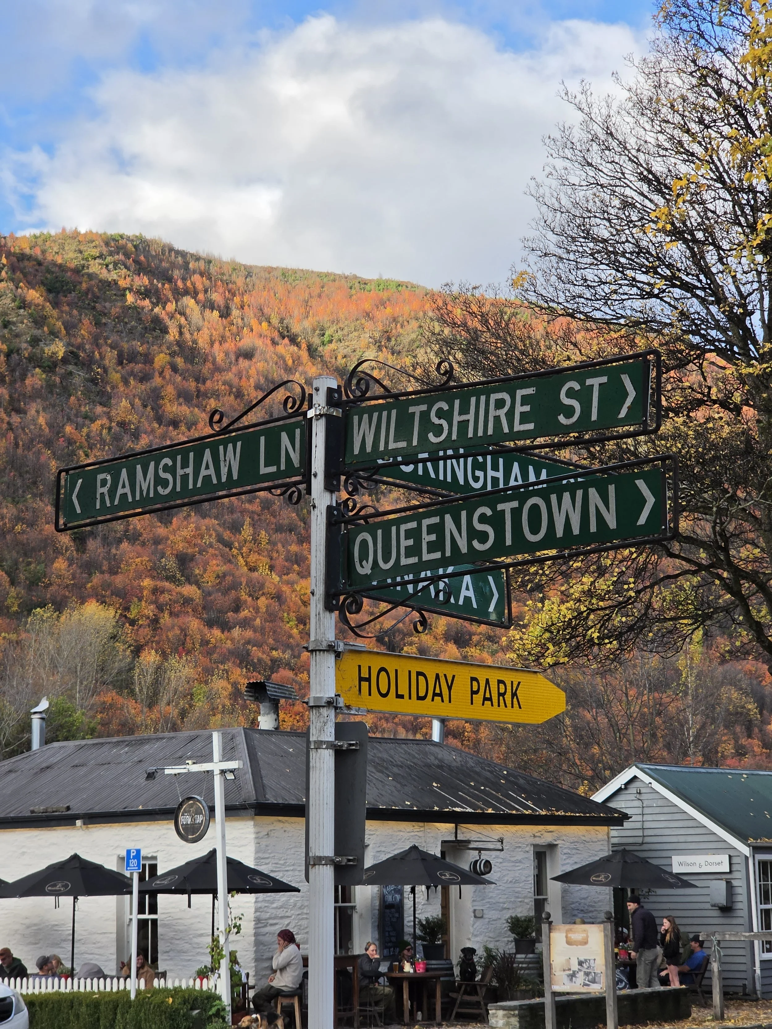 A street sign at Wilshire Street and Ramshaw Lane, with a background of fall-colored trees and a small outdoor dining area with umbrellas and people sitting.