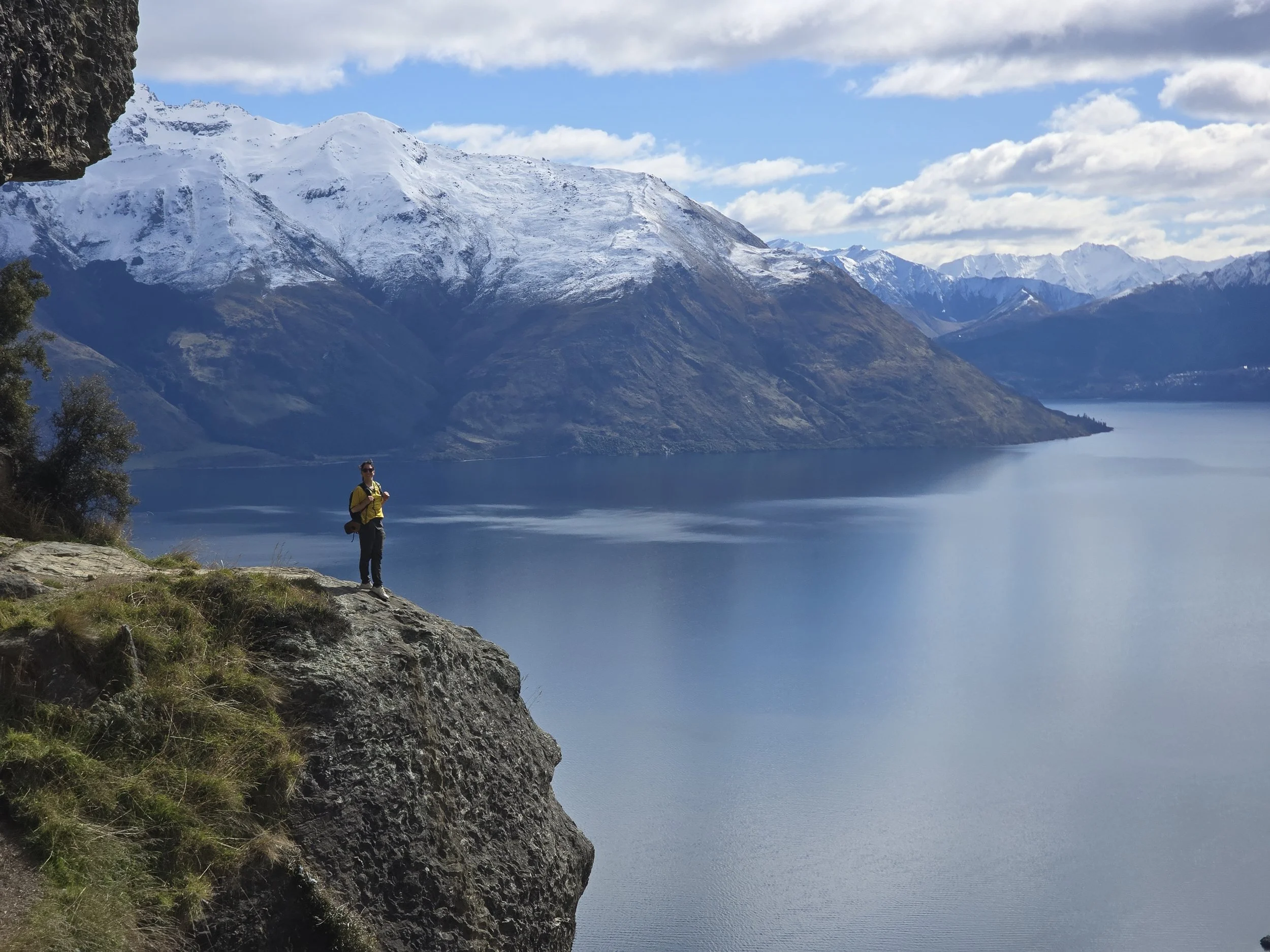 wye creek Person standing on a rocky ledge in front of a large body of water with snow-capped mountains in the background, under a partly cloudy sky.