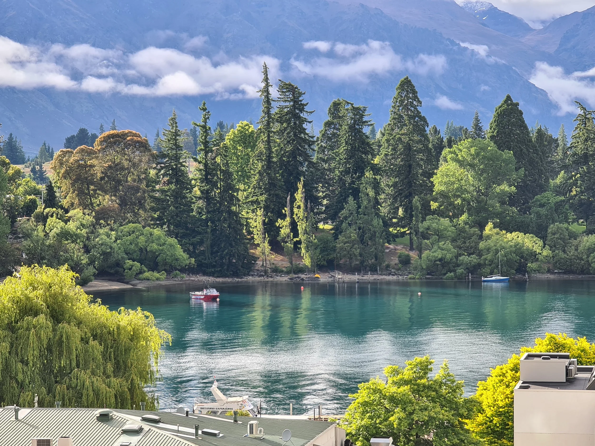 A scenic view of a lake with turquoise water, surrounded by lush green trees and plants, with boats floating on the water. In the background, there are mountains with clouds hovering around their peaks.