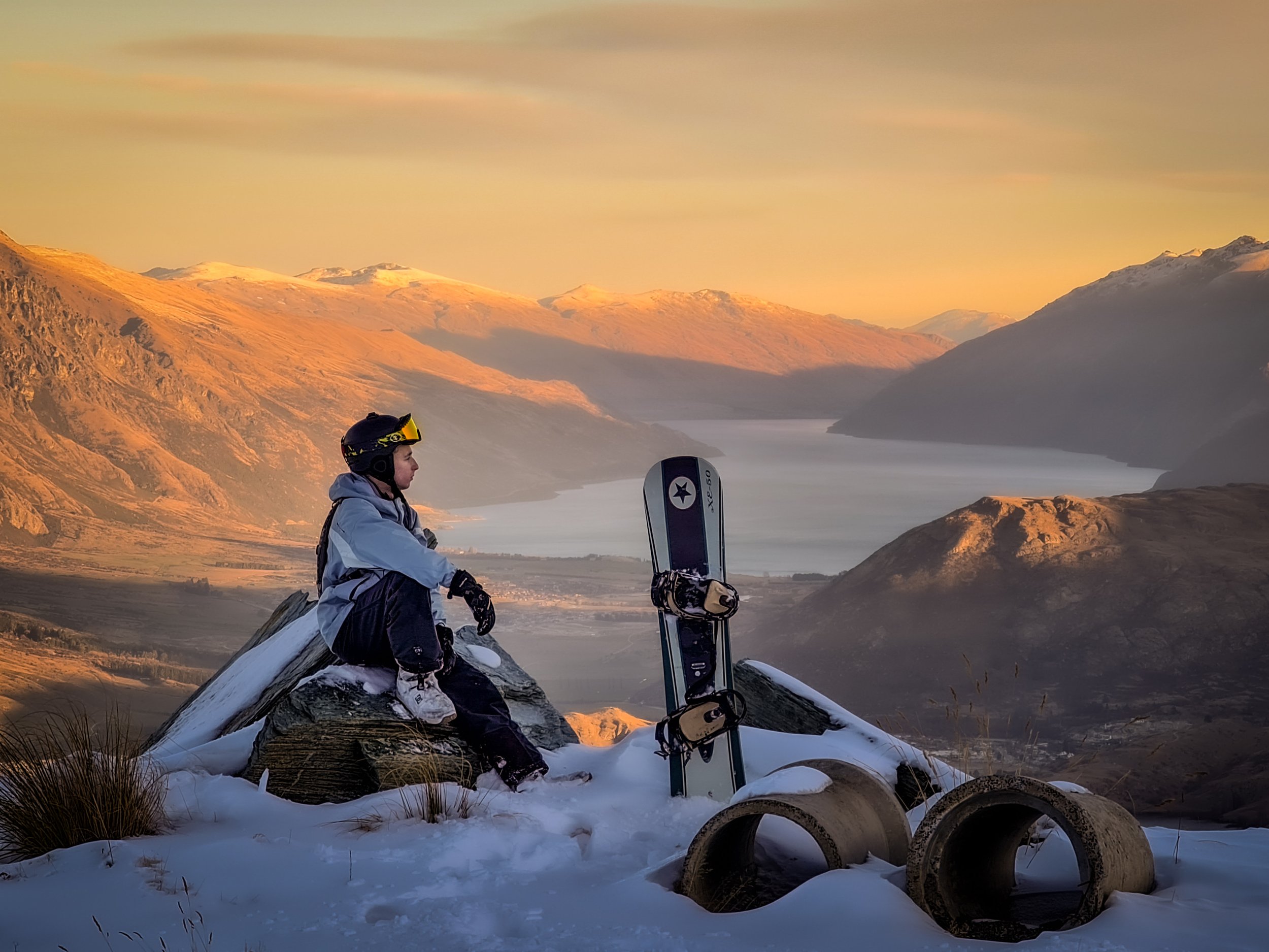 Person sitting on snow-covered rocks with skis nearby during sunset in mountainous landscape.