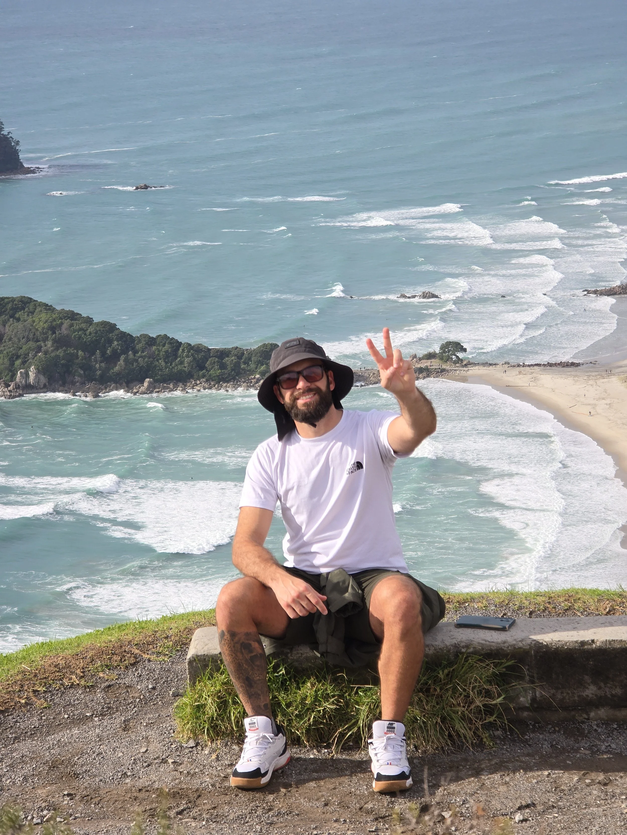 A man with a beard, wearing glasses, a hat, a white t-shirt, and shorts, is sitting on a rock near the coast, making a peace sign with his right hand. Behind him, there is a view of the ocean with waves hitting the shore and a beach area in the distance.