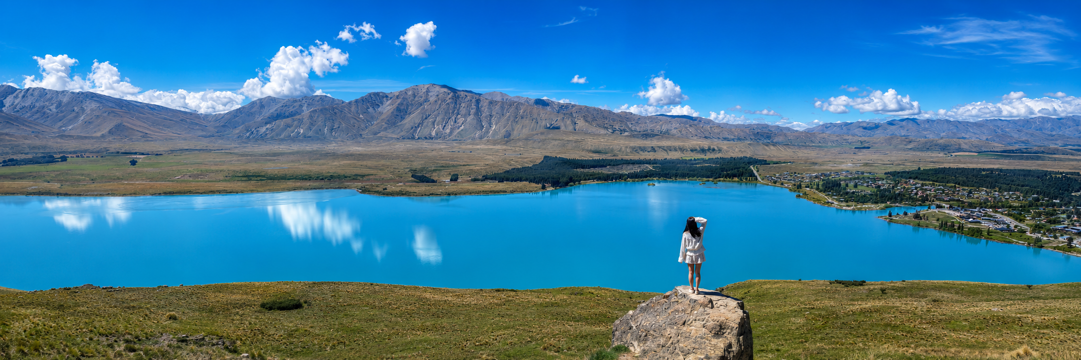 A woman standing on a rock overlooking a blue lake with mountains in the background under a partly cloudy sky.