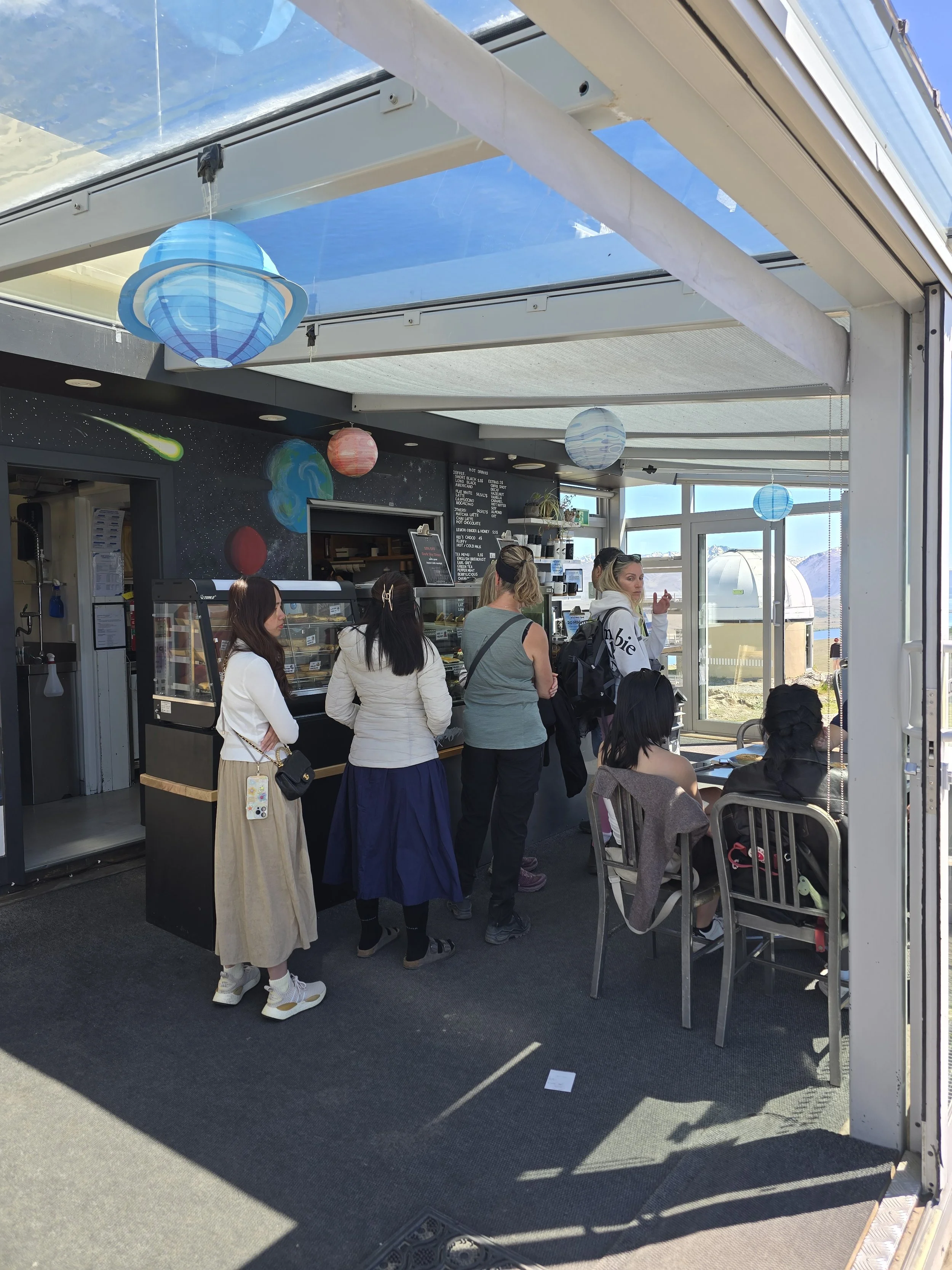 People waiting in line inside a food or coffee stand at an outdoor area with desk seating, solar panels, and planet-themed decorations hanging from the ceiling.