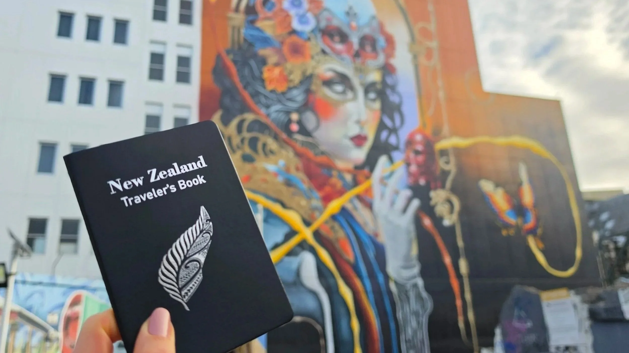 A person holding a black New Zealand traveler’s book with a fern logo in front of a colorful mural of a woman’s face with dark hair, red lips, and vibrant colors in an urban setting.