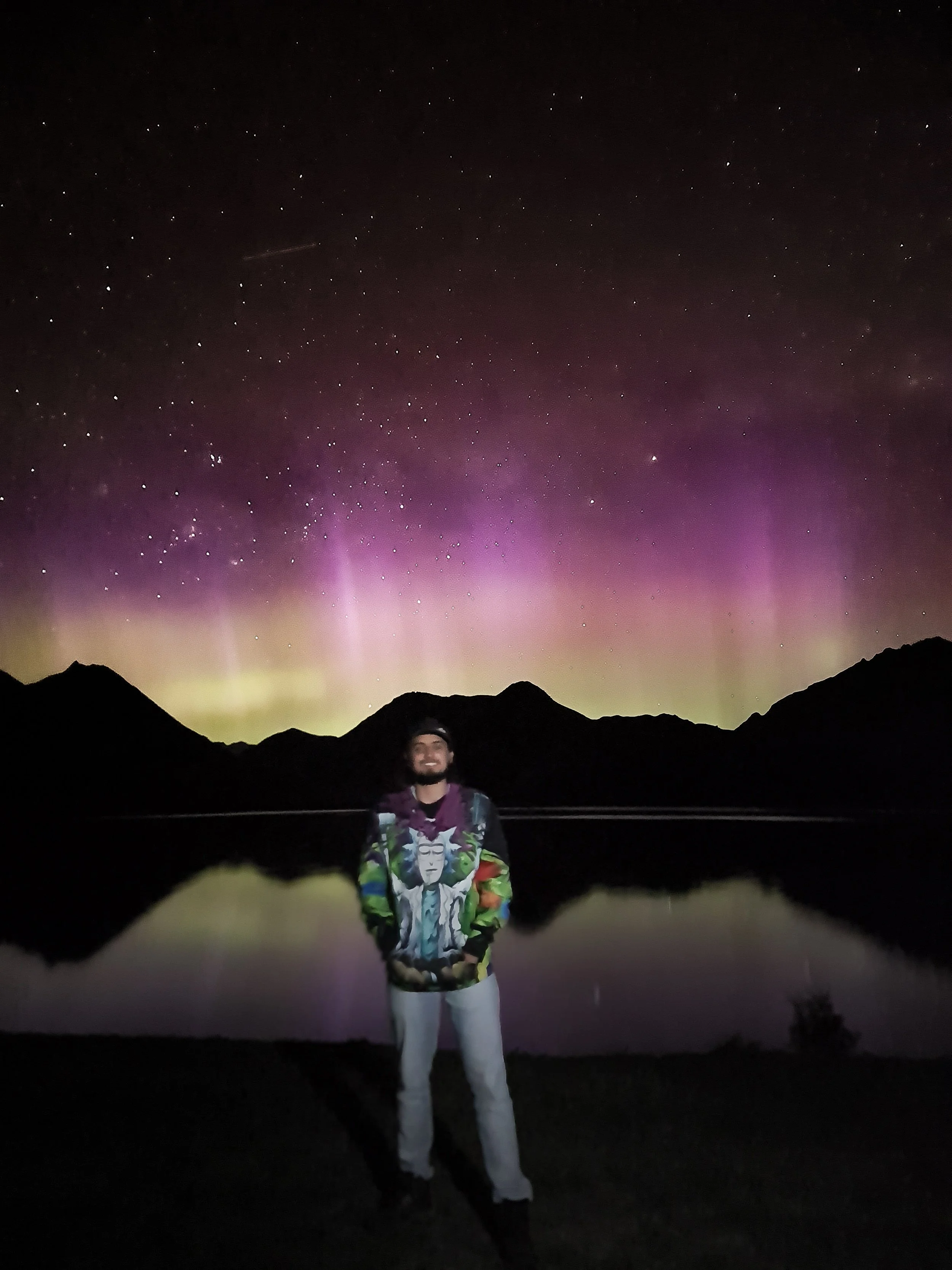 Person standing outdoors at night with mountain silhouette in background and the Northern Lights visible in the sky.