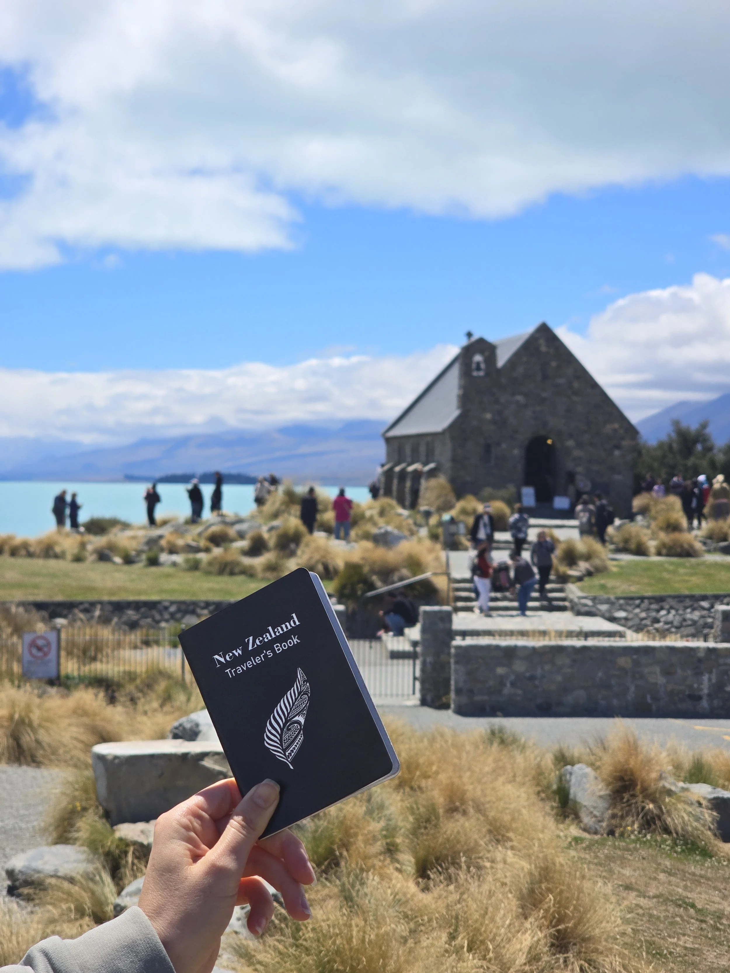 Person holding a brochure titled 'New Zealand Traveler's Book' with a historic stone church and visitors gathered outside, set in a scenic landscape with cloudy sky and mountains in the background.