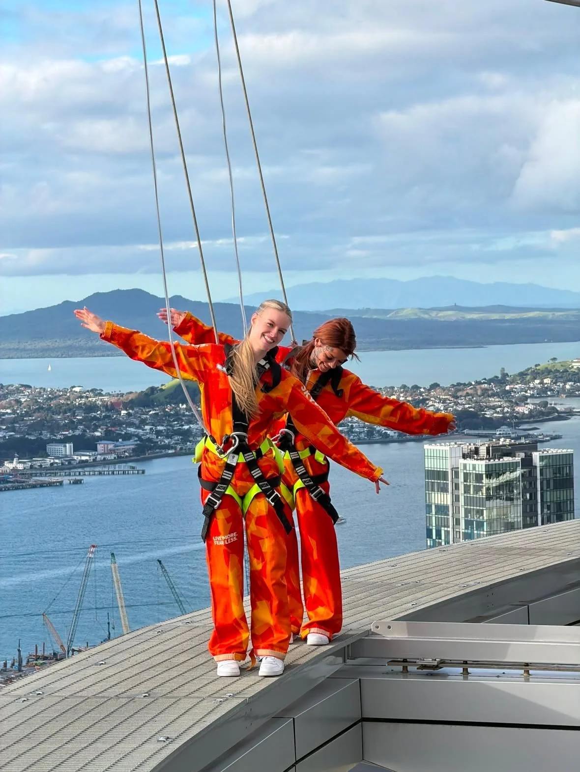 Two women in orange jumpsuits are standing on the edge of a rooftop with a city and water view behind them. They are wearing harnesses and are strapped in for a safety activity, smiling and posing with arms outstretched.