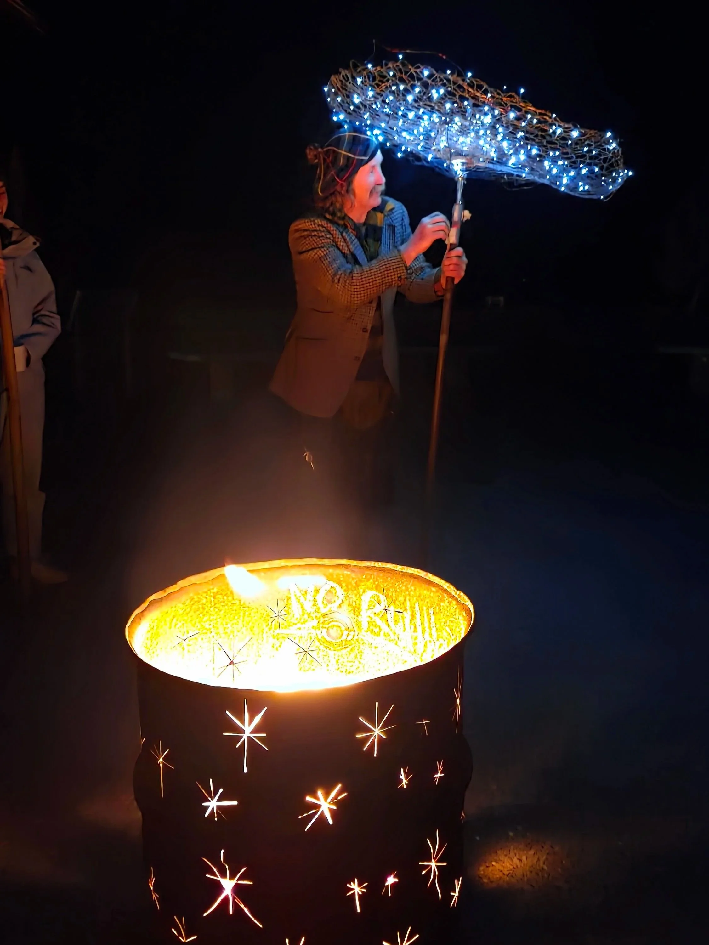 Person holding an umbrella decorated with blue LED lights participating in a nighttime fireworks display, with sparks and glowing fireworks in the foreground.