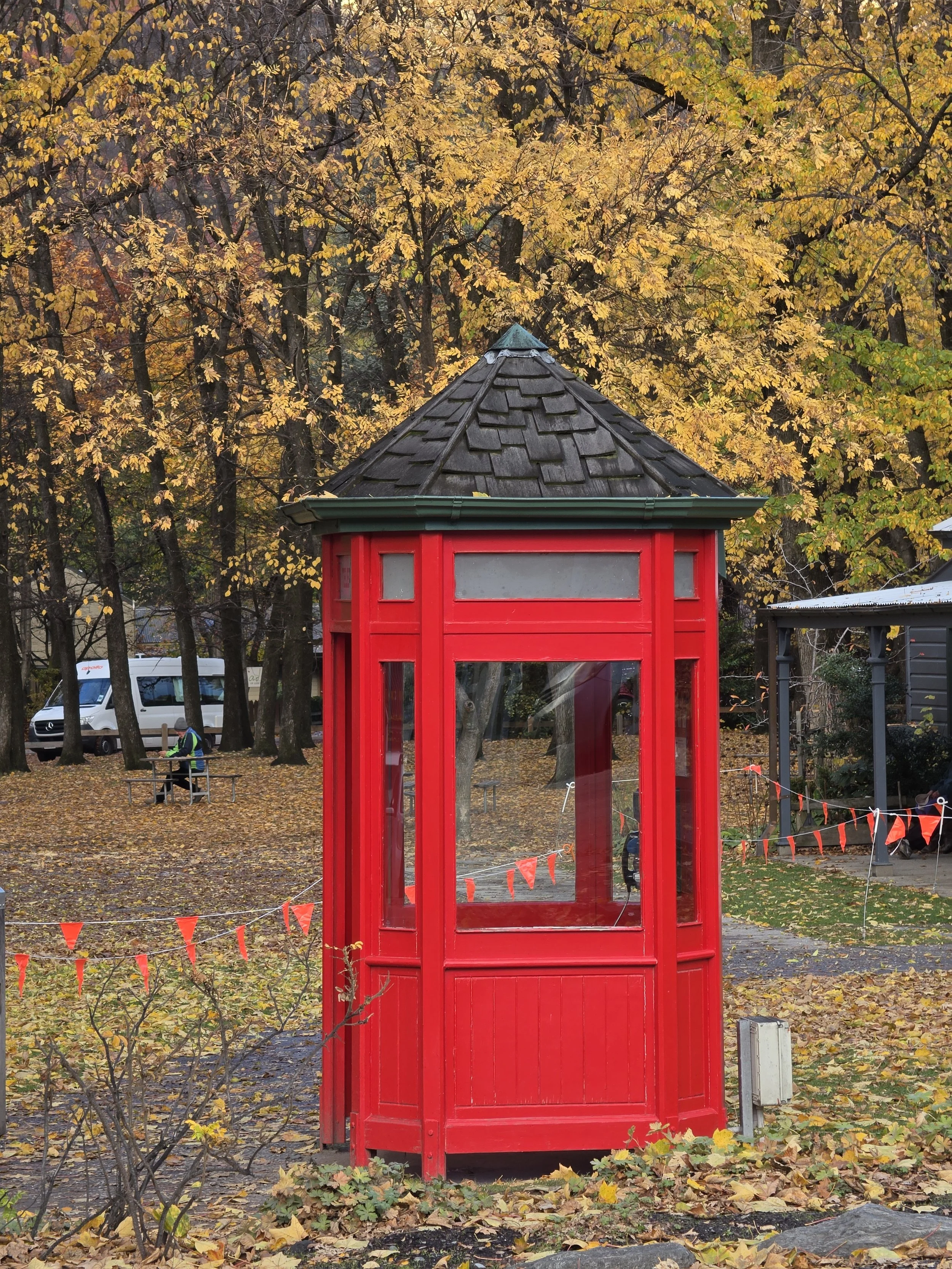 A red telephone booth standing in a park with autumn leaves on the ground and trees with yellow and orange foliage in the background.