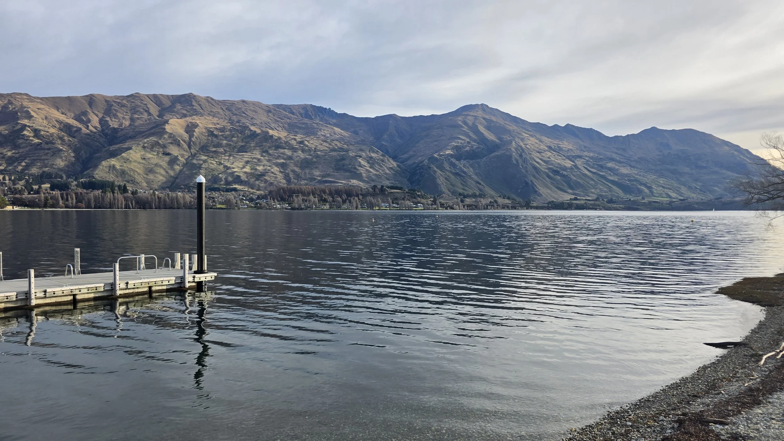 A serene lakeside scene with a pier extending into the water, calm water reflecting the cloudy sky, and mountains in the background.