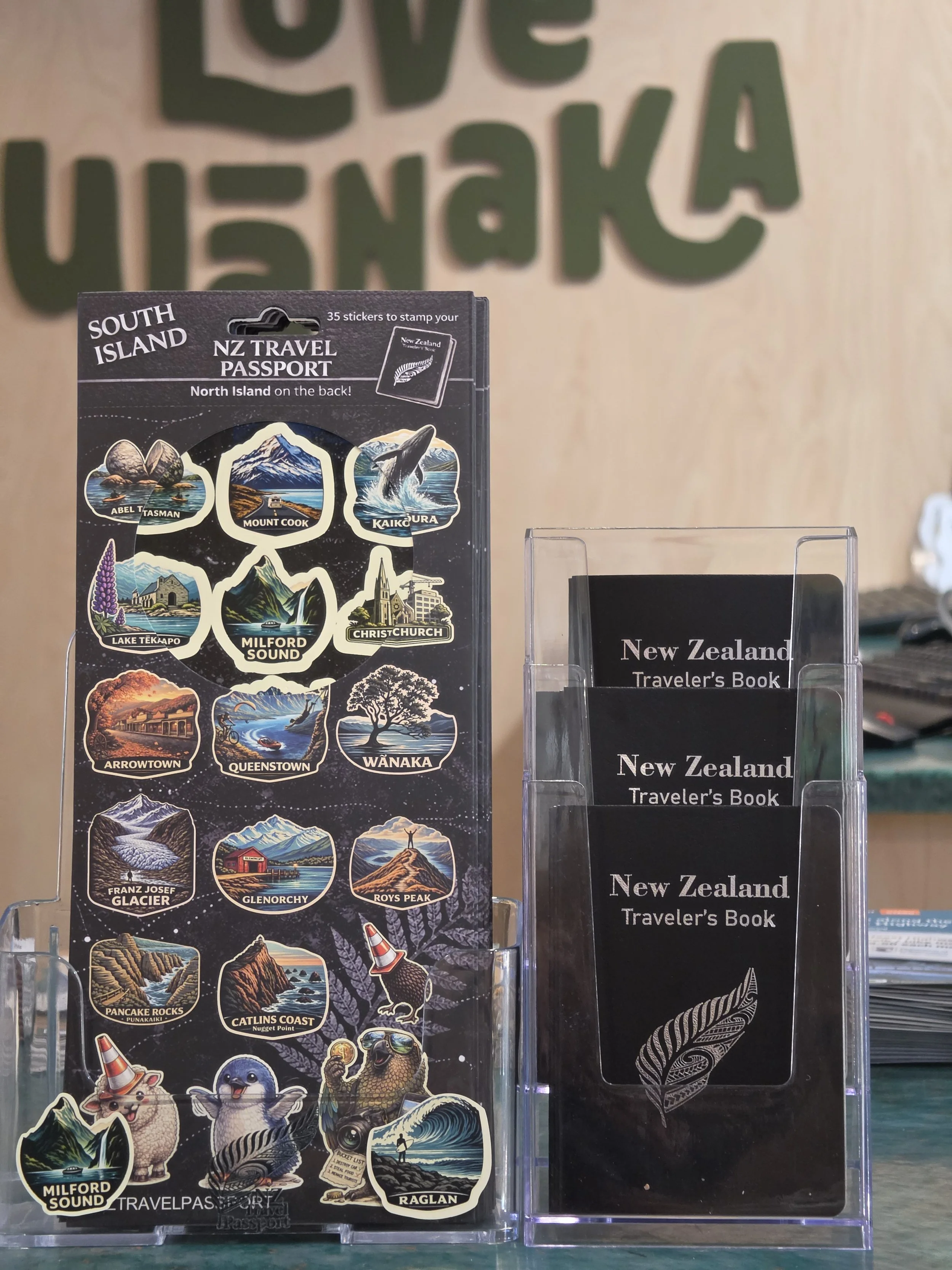 A display of New Zealand-themed travel stickers and brochures. The stickers feature various landmarks and animals, including Mount Cook, Kaikoura, Milford Sound, and the Wānaka tree, as well as animals like penguins and a lamb. To the right, there are brochures titled 'New Zealand Traveler's Book' in a transparent holder.
