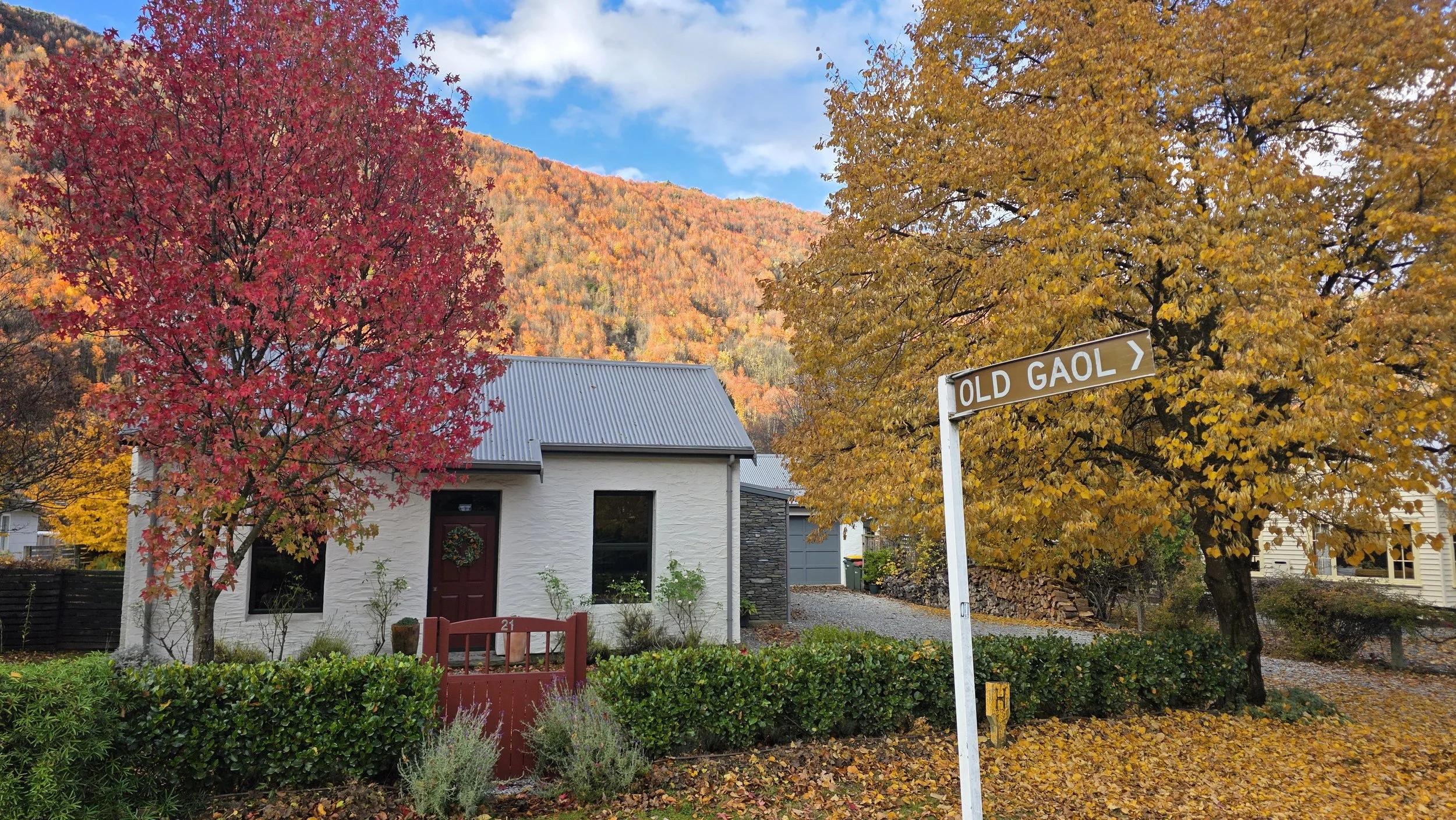 A white house with a dark red front door decorated with a wreath, surrounded by autumn trees with red and yellow leaves. In the background, there are mountains with colorful fall foliage, and a street sign that reads 'OLD GAOL'.