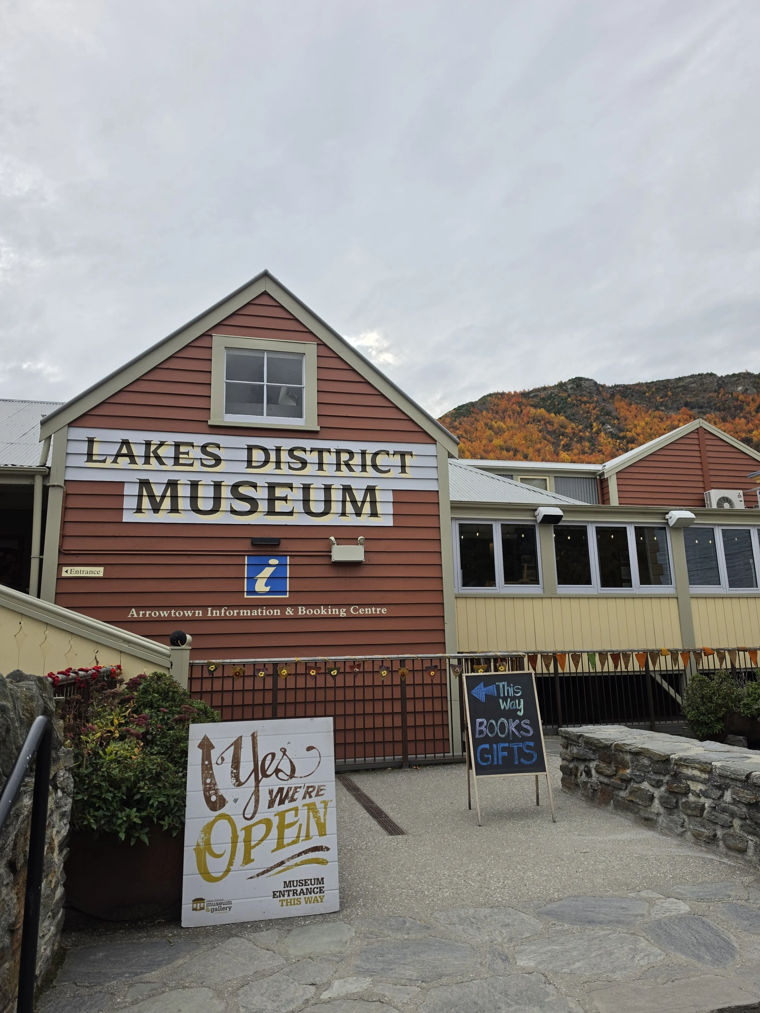 Entrance of the Lakes District Museum with a large sign saying 'Yes We're Open' and smaller signs with directions and information, surrounded by plants and a stone wall, with a building and fall foliage mountain in the background.