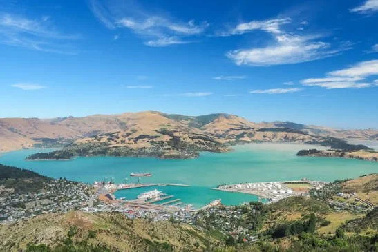 A panoramic view of a harbor with boats, surrounded by hills and a town, under a blue sky with scattered clouds.