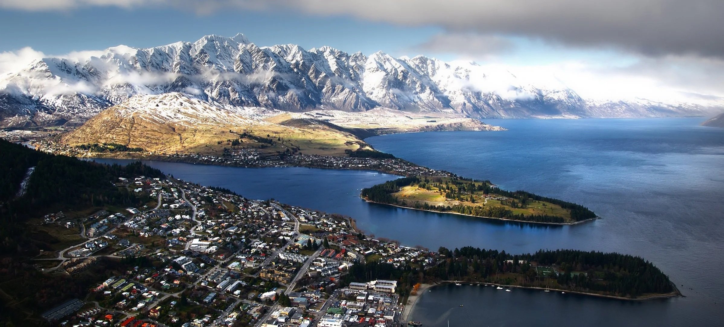 Aerial view of a town beside a large body of water, surrounded by mountains with snow-covered peaks.