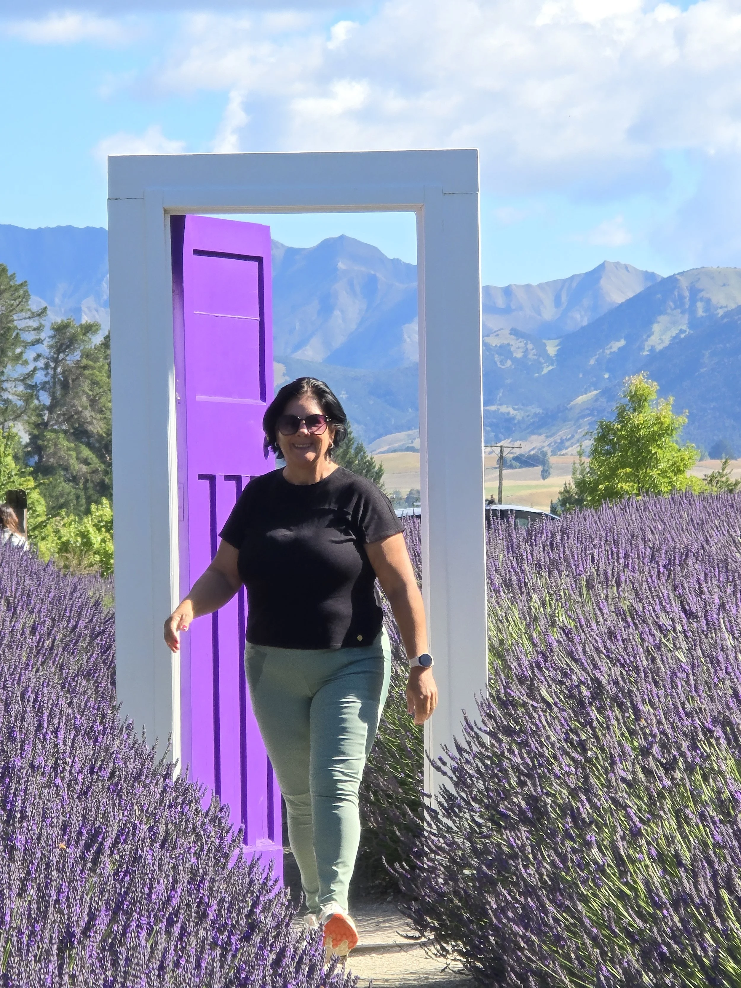 A woman wearing sunglasses, a black t-shirt, beige pants, and orange shoes is walking through a field of purple lavender flowers. She is smiling and holding her right arm out. Behind her, a large purple door is framed by a white square structure. In the background, there are mountains under a partly cloudy sky.