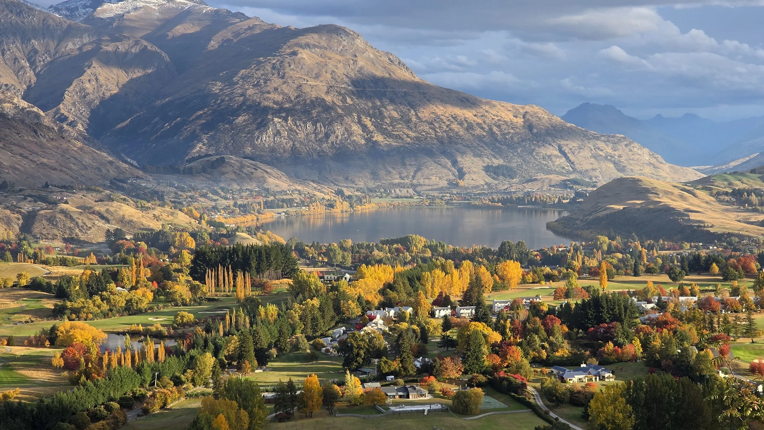 A scenic landscape of a valley with colorful autumn trees, a lake, and mountain ranges in the background under a cloudy sky.