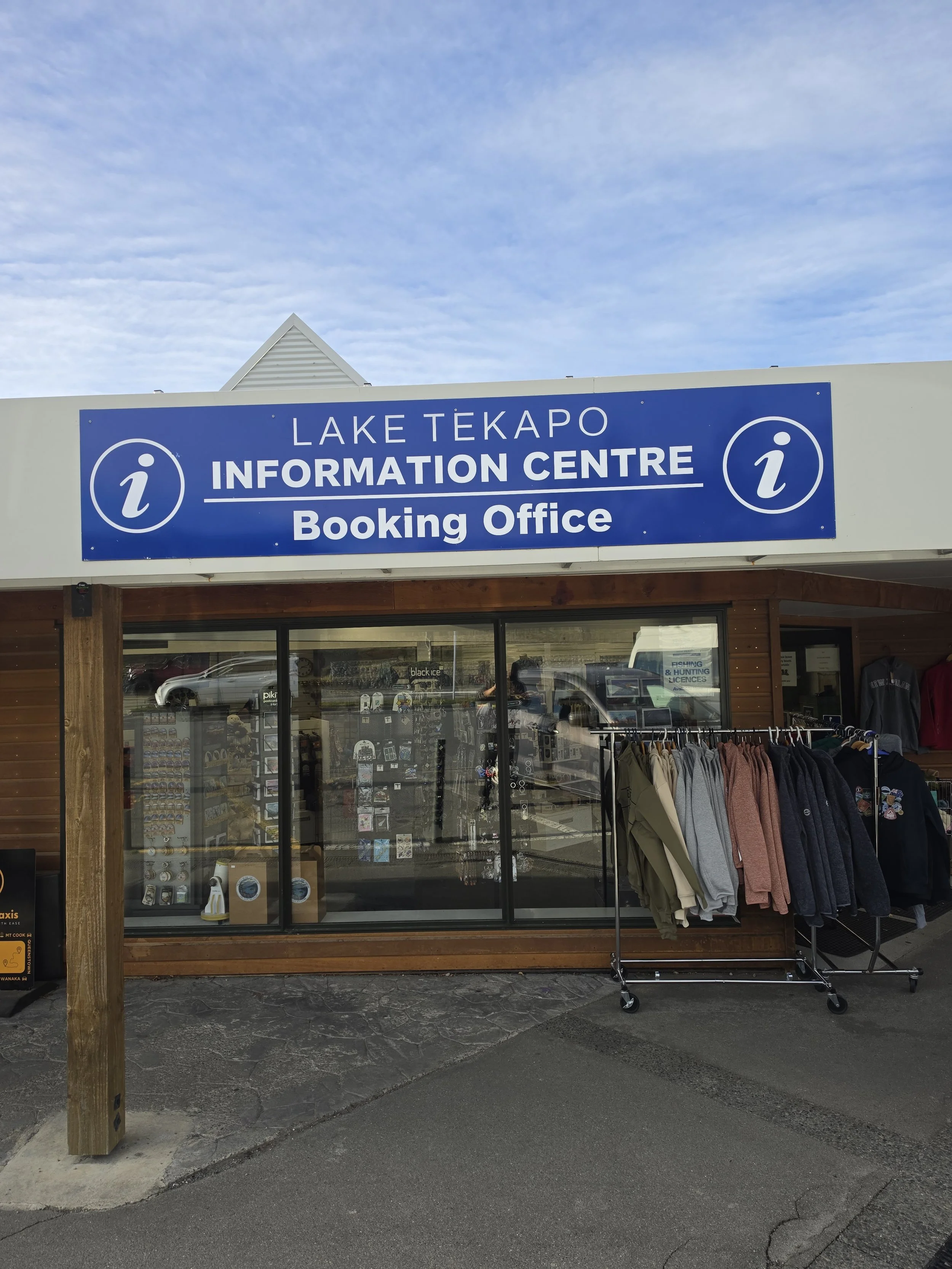 Building with sign reading 'Lake Tekapo Information Centre Booking Office' and clothing racks outside, with cars and a cloudy sky in the background.