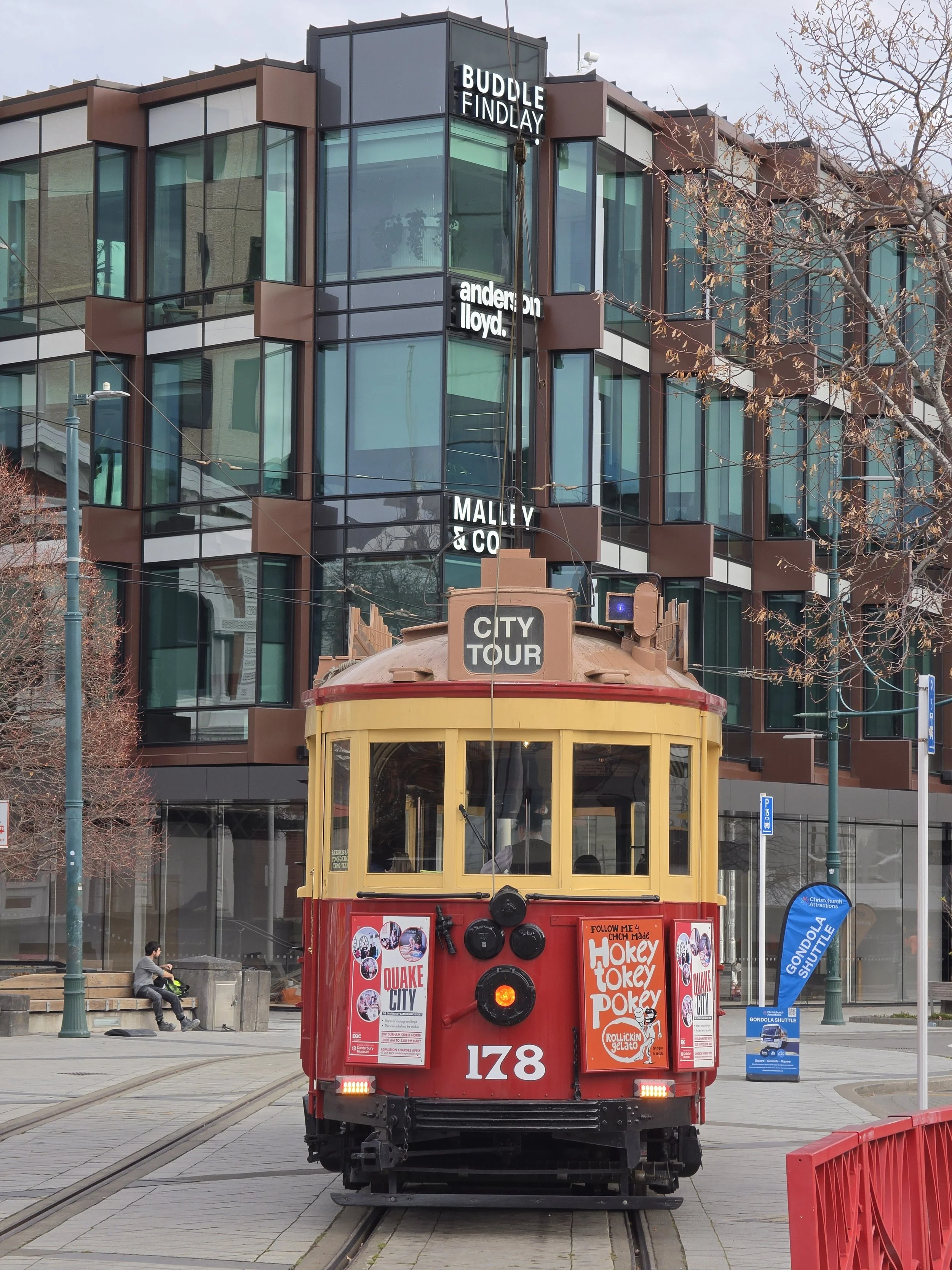 A red vintage trolley car on tram tracks in an urban setting with a modern glass building in the background. The tram is decorated with posters and has the number 178 displayed on the front.