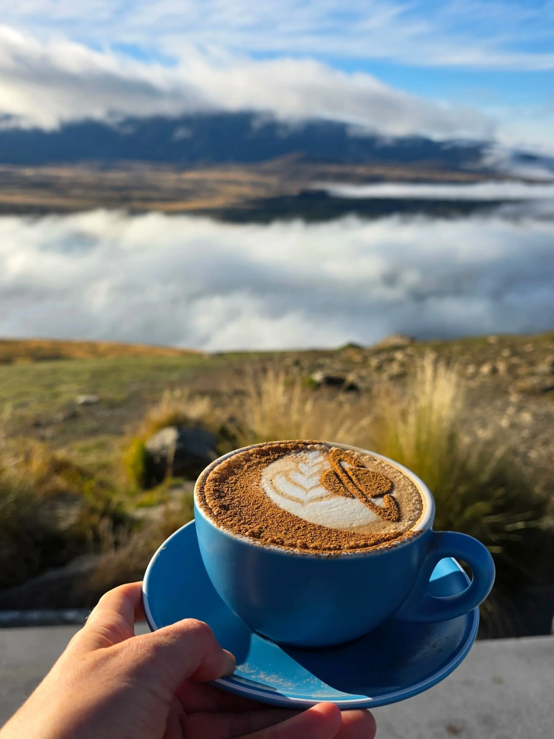 A hand holding a blue cup of coffee with latte art, set against a landscape of mountains, clouds, and sky in the background.