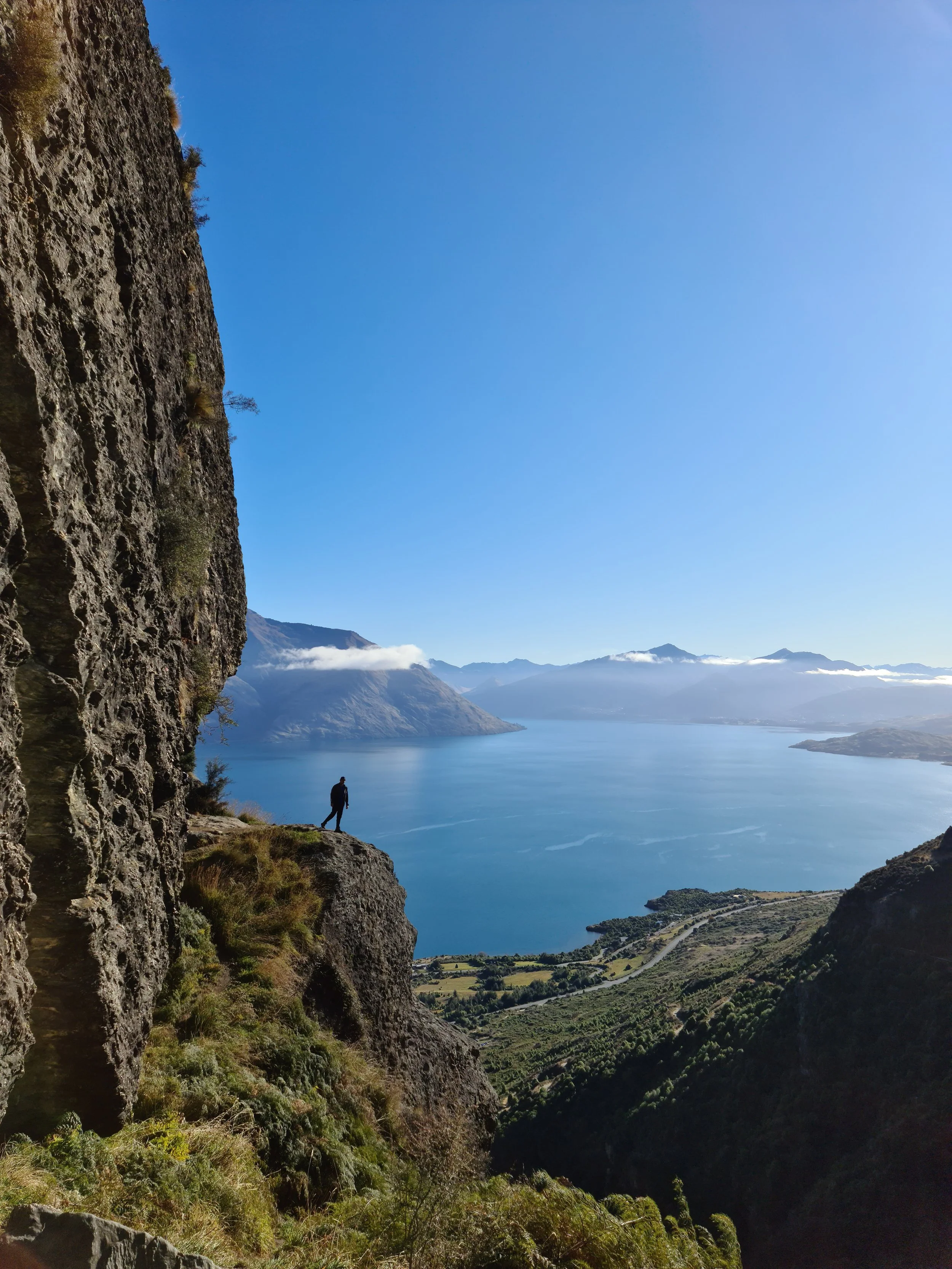 wye creek  A person walking on a rocky trail beside a mountain cliff with a large lake and distant mountains in the background under a clear blue sky.