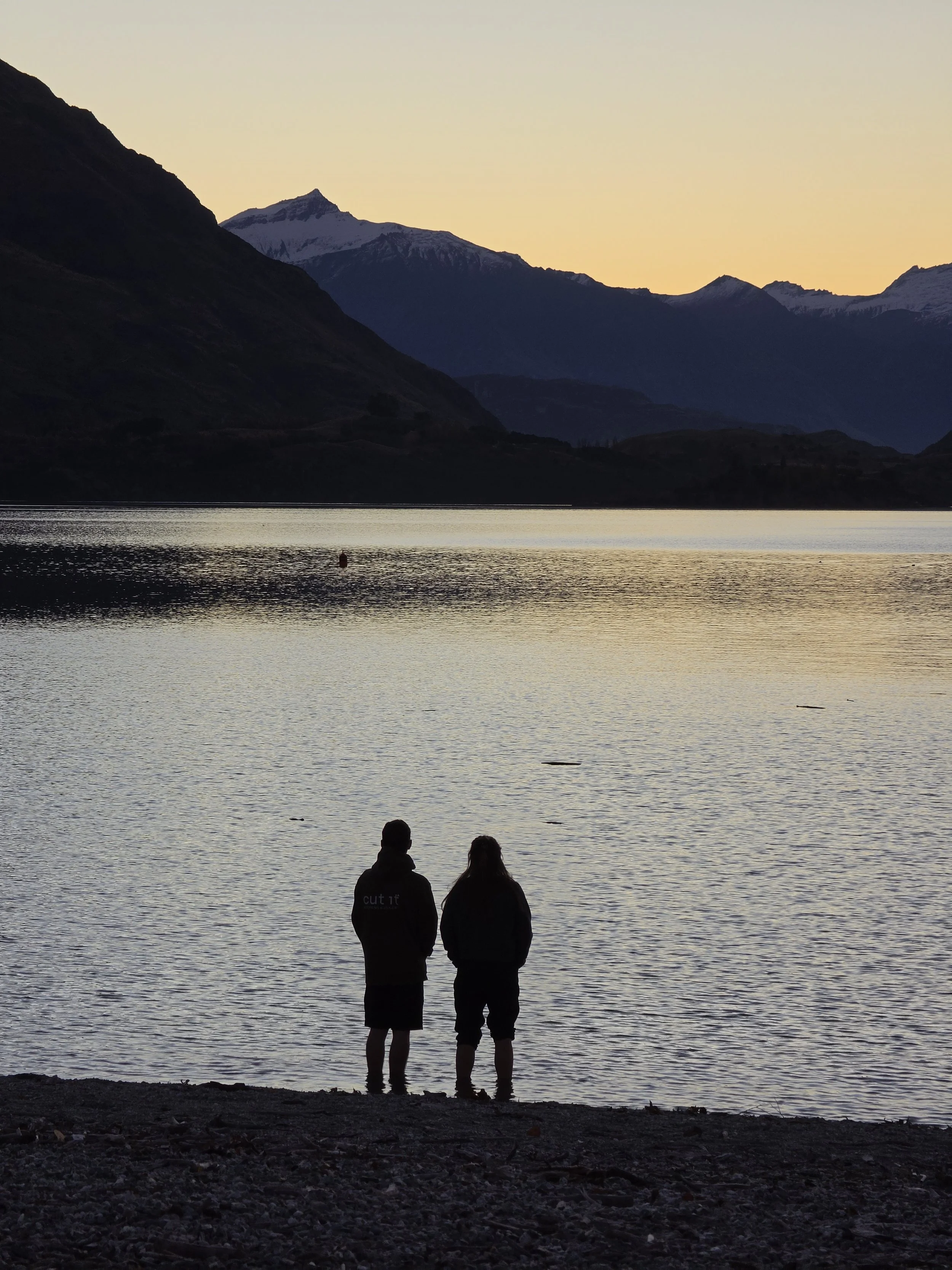 Two people standing at the edge of a body of water during sunset or sunrise, with mountains in the background and the sky lit with soft yellow light.