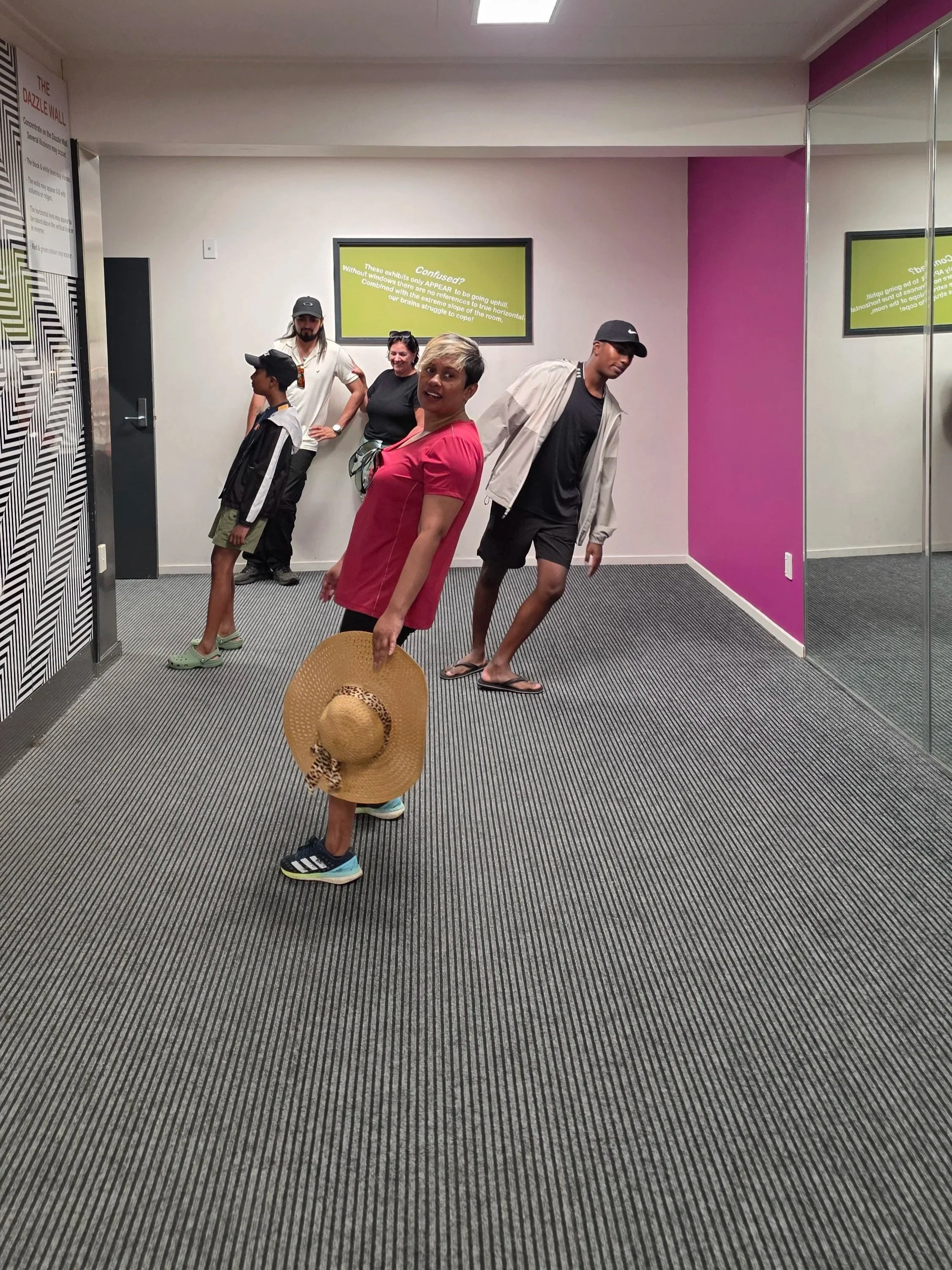 People standing inside a room with pink and white walls, some looking at the camera, one holding a wide-brimmed hat, others in casual attire, with a display screen on the wall behind them.