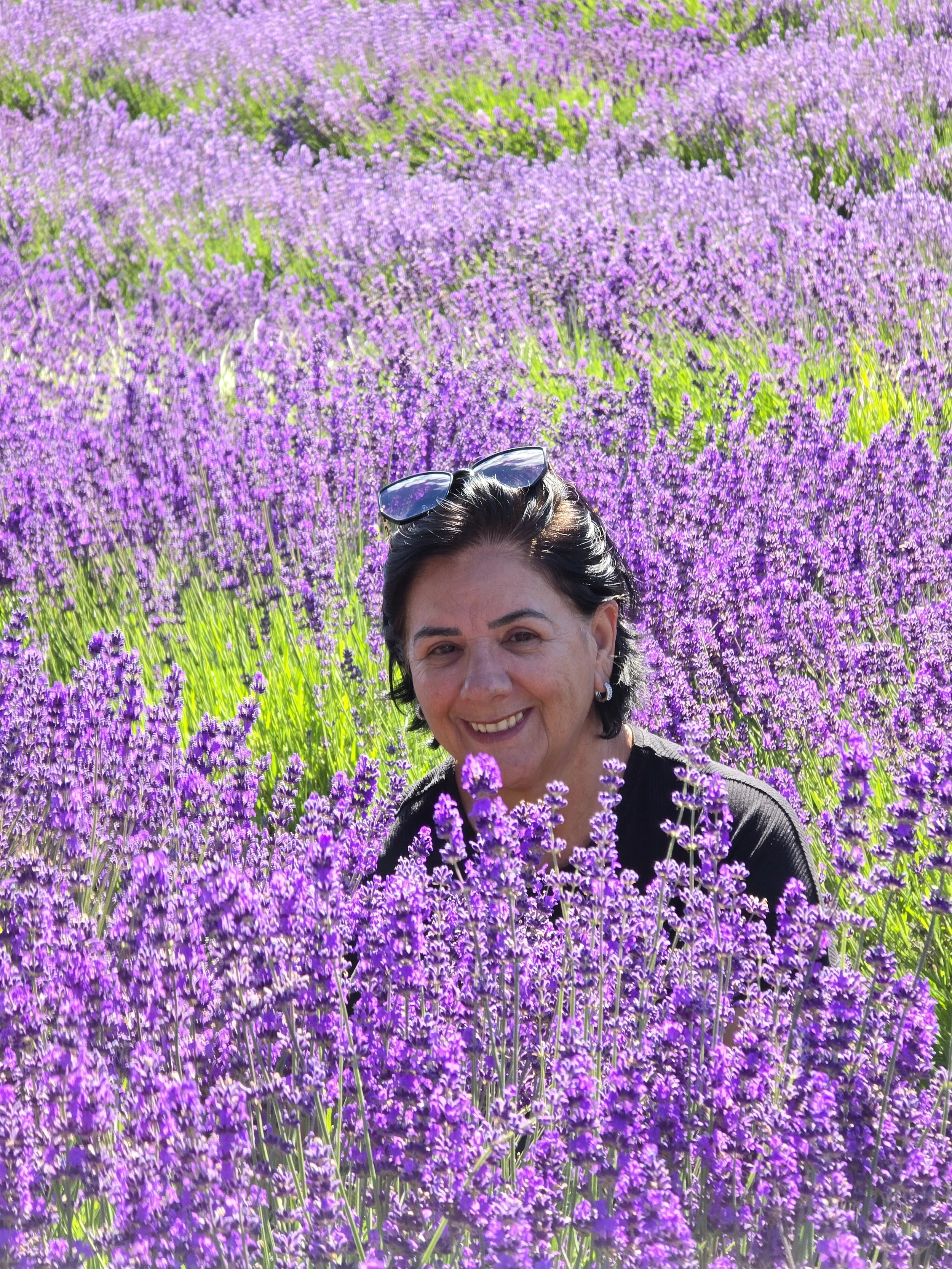 A woman with dark hair and sunglasses on her head, smiling among a field of purple lavender flowers.