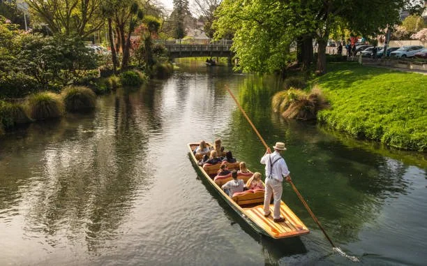 A guide in a white shirt and hat steers a small boat carrying several passengers along a calm river surrounded by green trees and grassy banks.