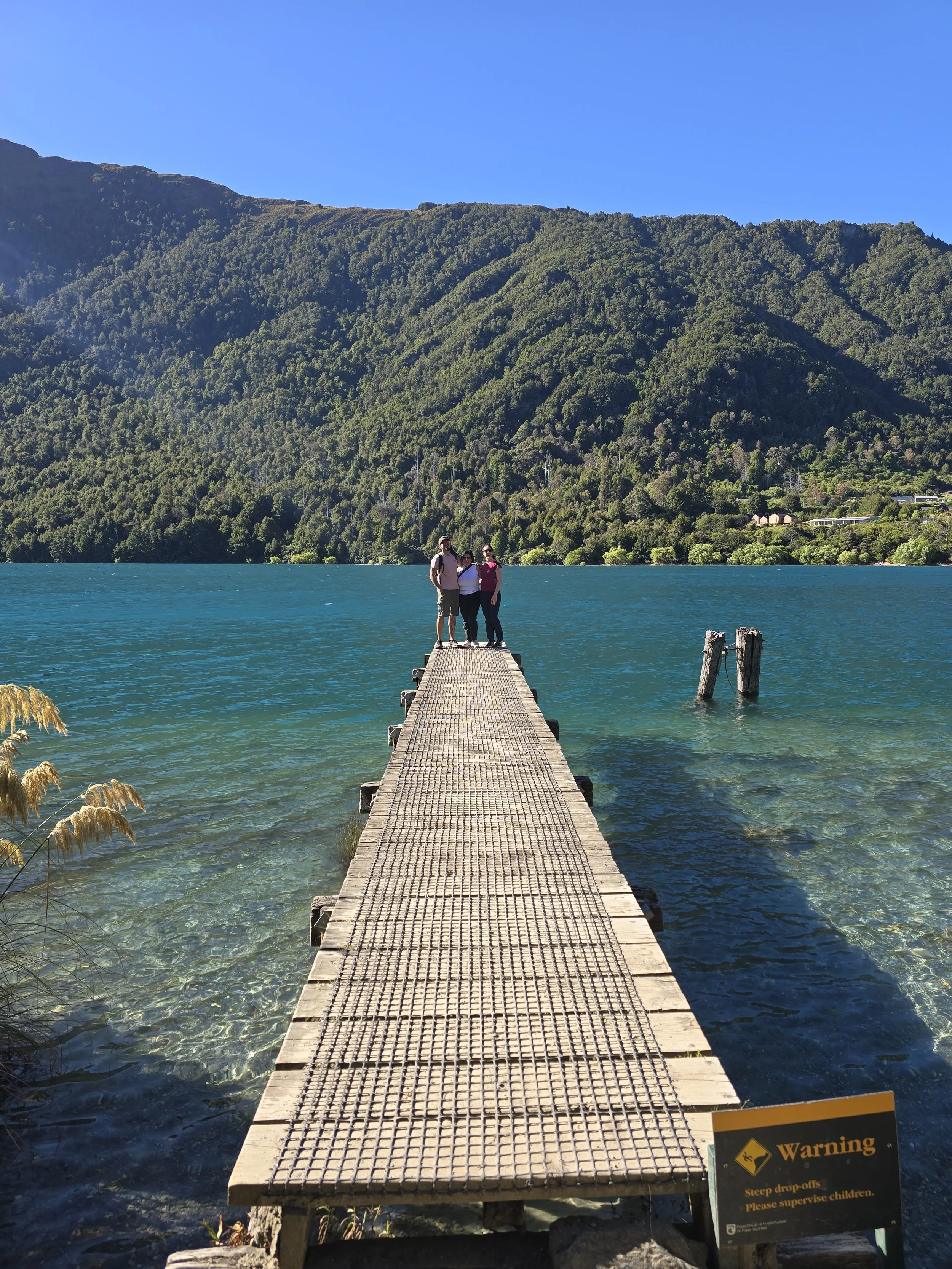 A narrow wooden pier extending into a lake with three people standing at the end, surrounded by lush green mountains and a clear blue sky.