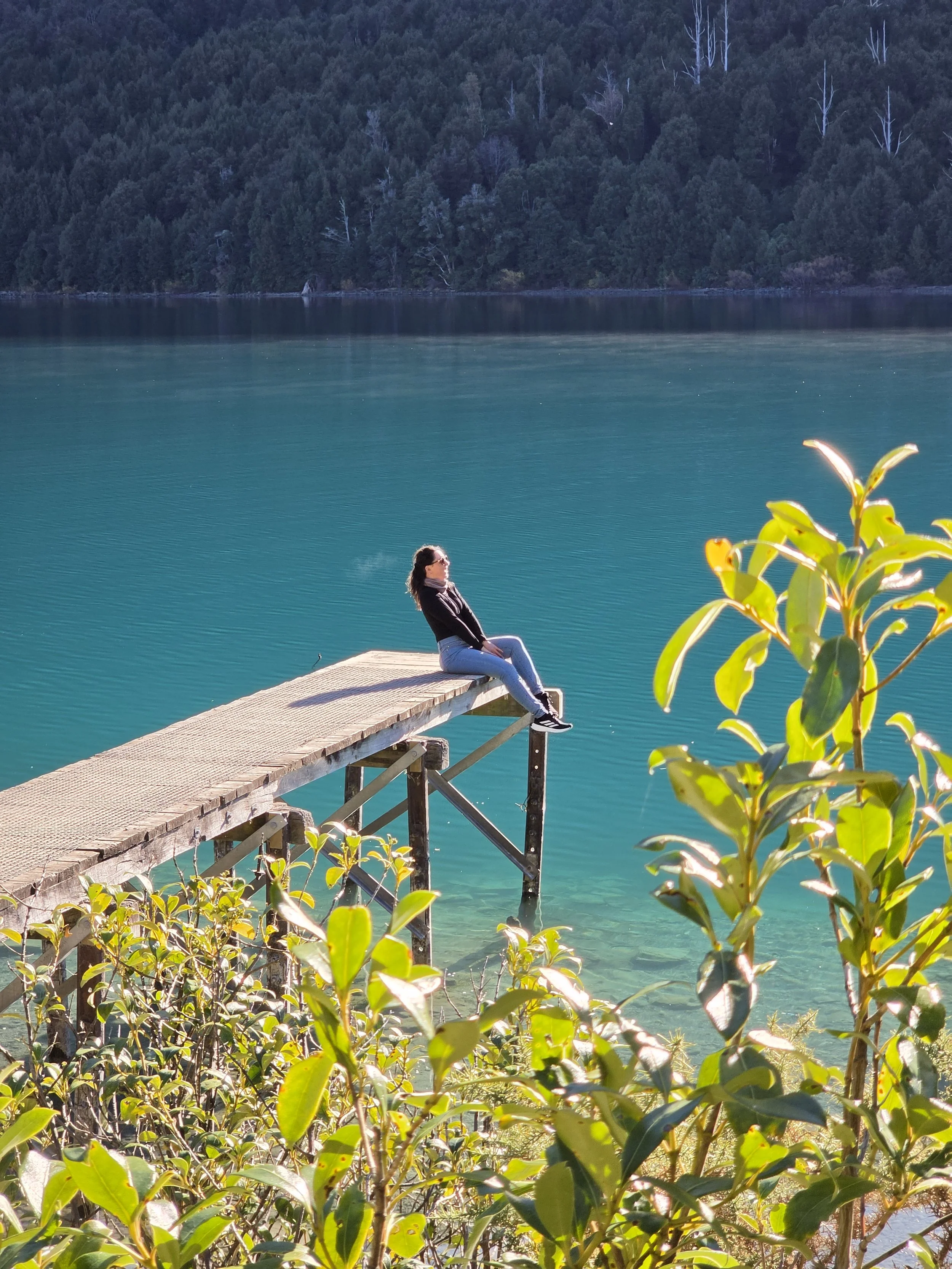 A woman sitting on a dock by a lake, surrounded by green foliage with a forested hillside in the background.